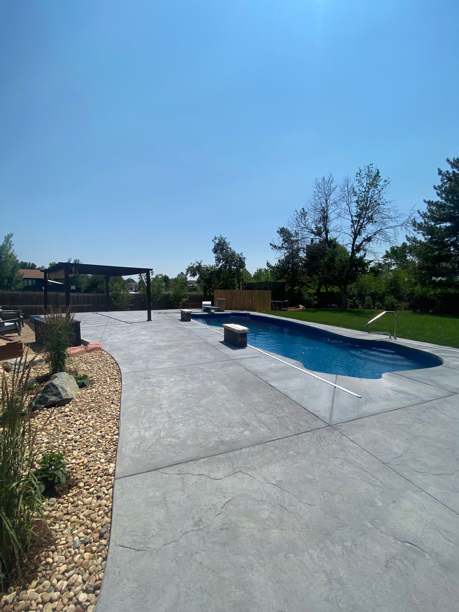A backyard pool with a concrete patio, landscaping, and a blue sky overhead.