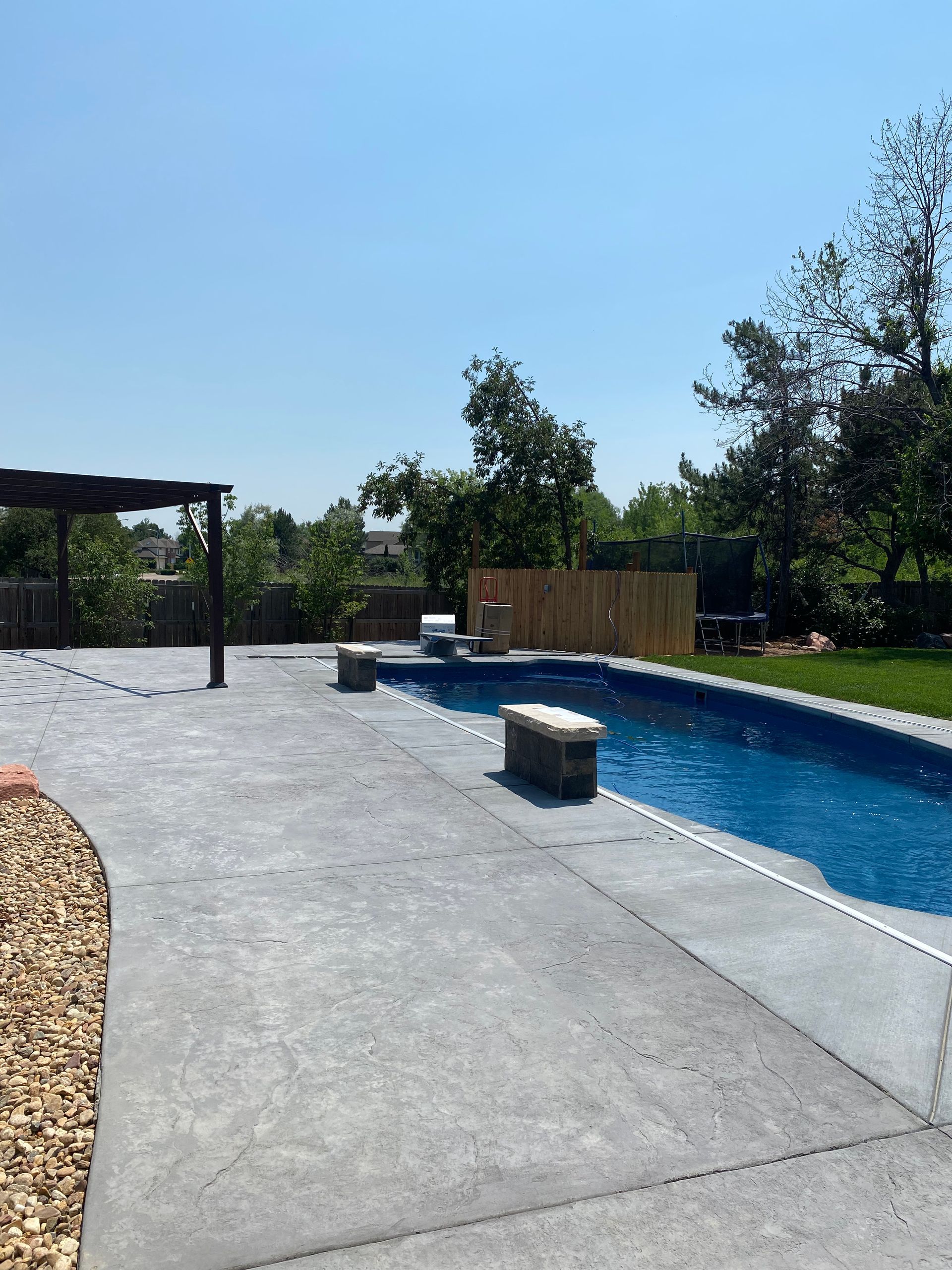 Concrete patio with a pool, pergola, and wooden fence on a sunny day.