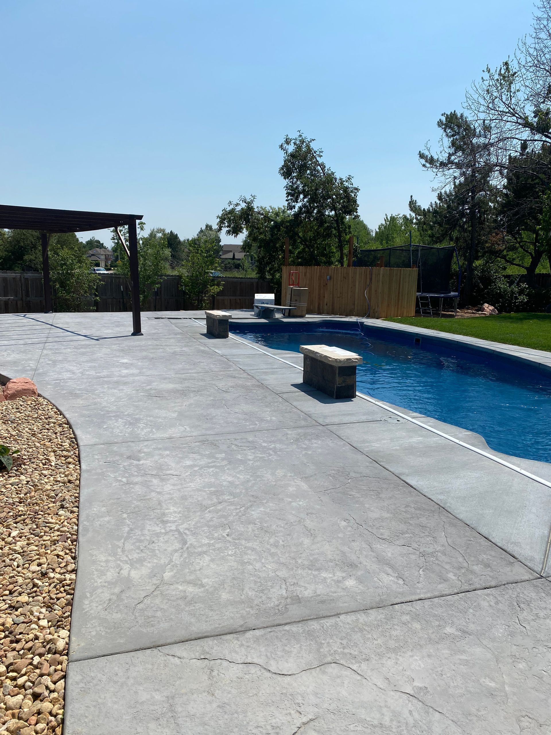 Backyard with a pool, concrete patio, pergola, and landscaping on a sunny day.