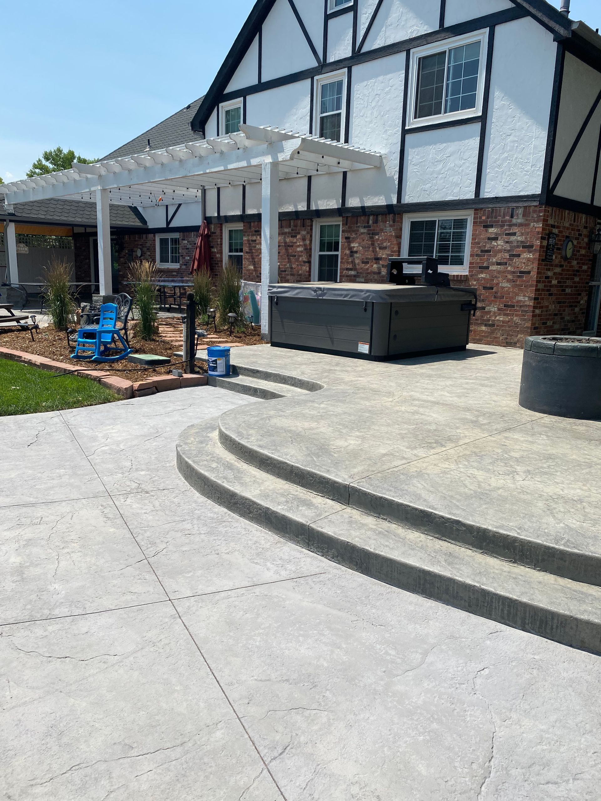 Concrete patio with hot tub, pergola, and brick house. Blue chair on grass.