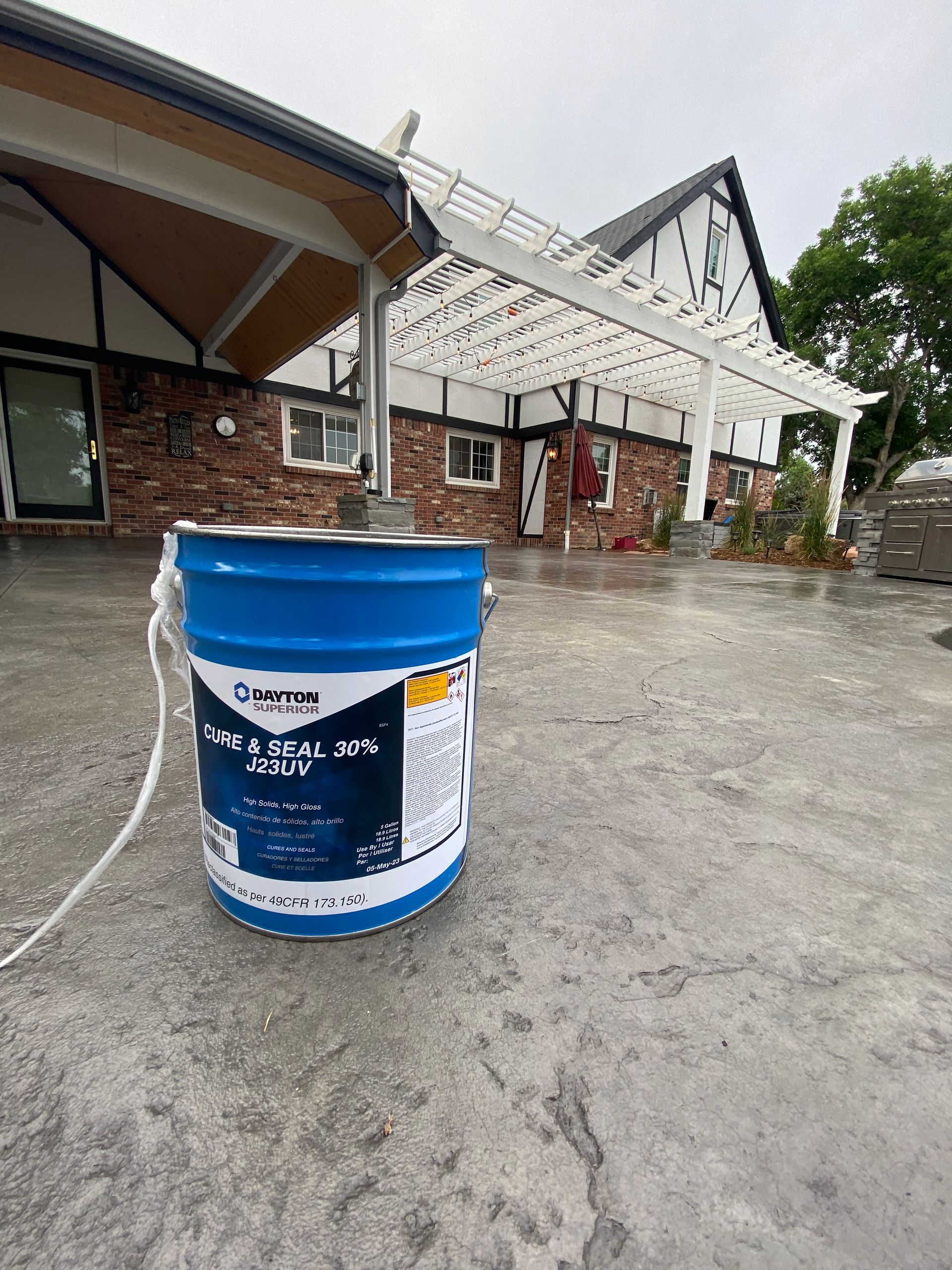 Blue paint bucket with a mixer on a concrete patio, house in the background.