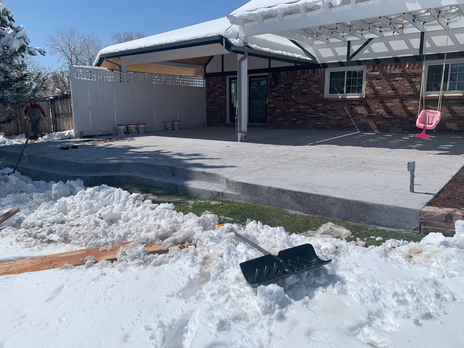Snow-covered backyard with a concrete patio, a pergola, and a pink swing. A shovel is in the snow.