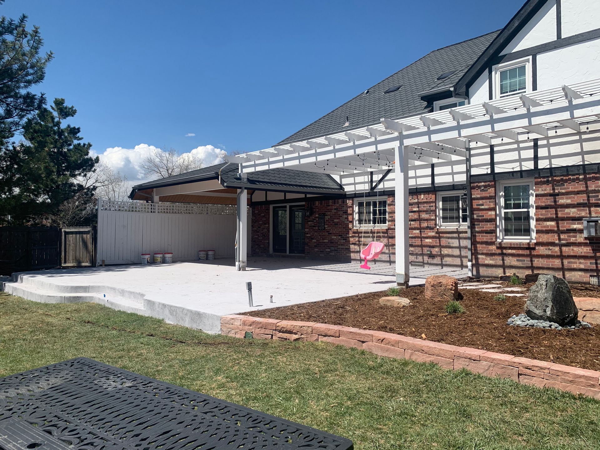 Backyard patio with pergola, house with brick and white trim, concrete patio, blue sky.