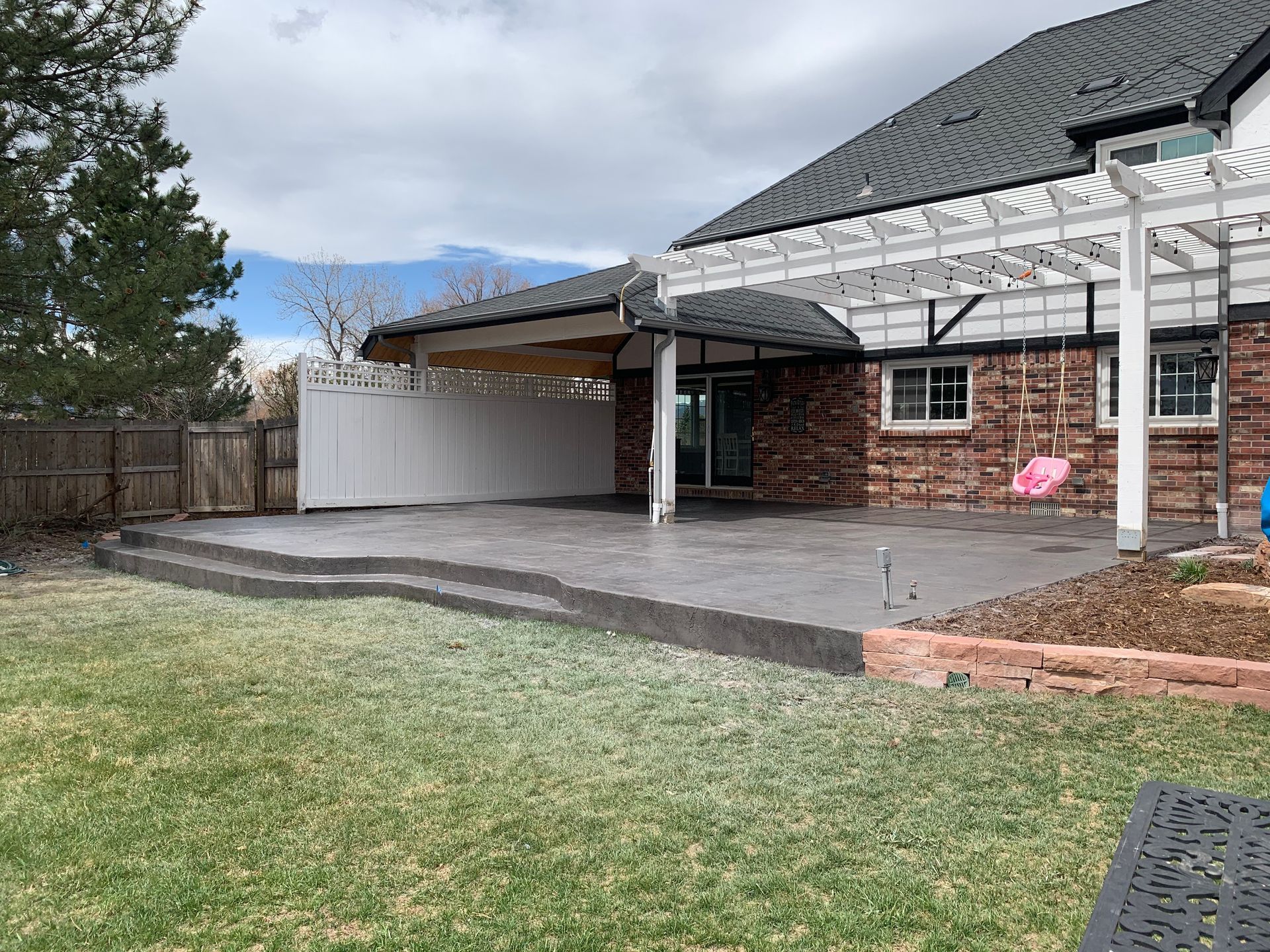 Backyard patio with steps, concrete, covered section, and house. Green grass and fence surround it.