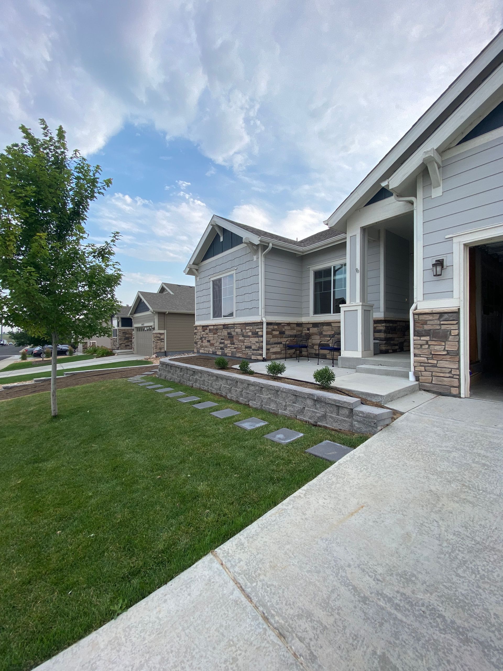 Gray house with a stone and gray facade, a cement walkway, and a green lawn under a cloudy sky.