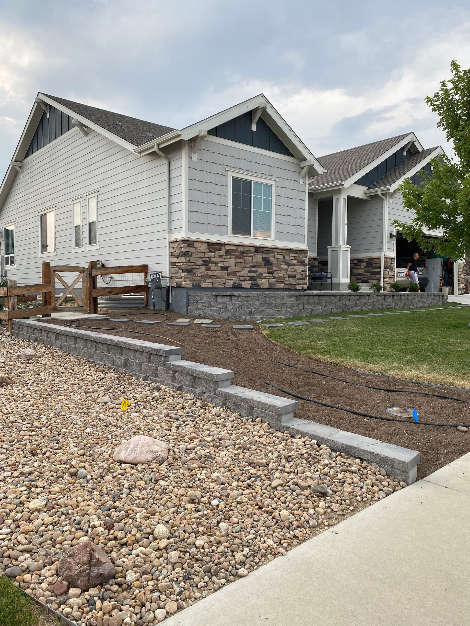 Light blue house with stone accents, brown gravel landscaping, gabion walls, cloudy sky.