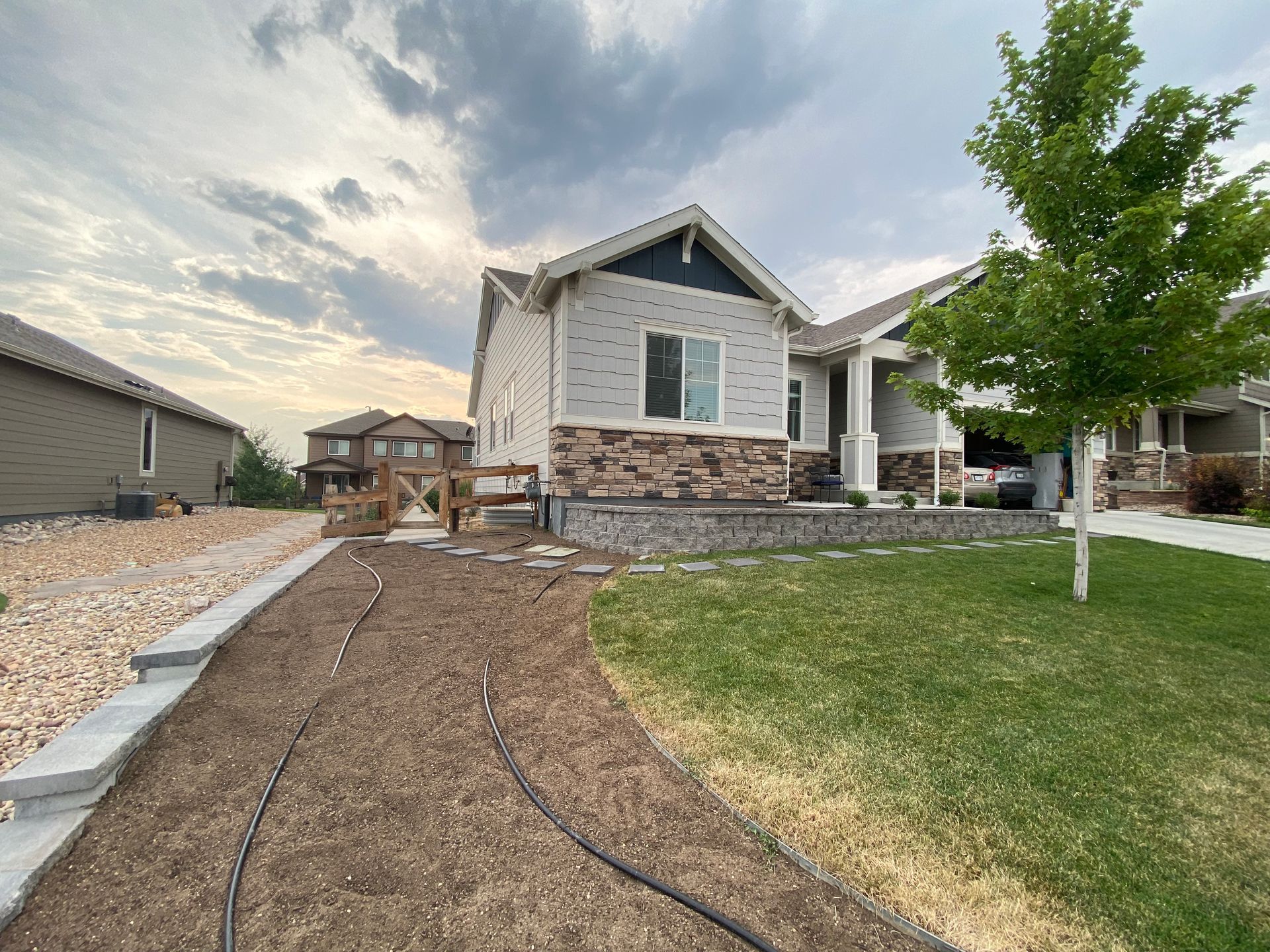 A house with light brick and stone facade, pathway with irrigation, and lawn. Cloudy sky.