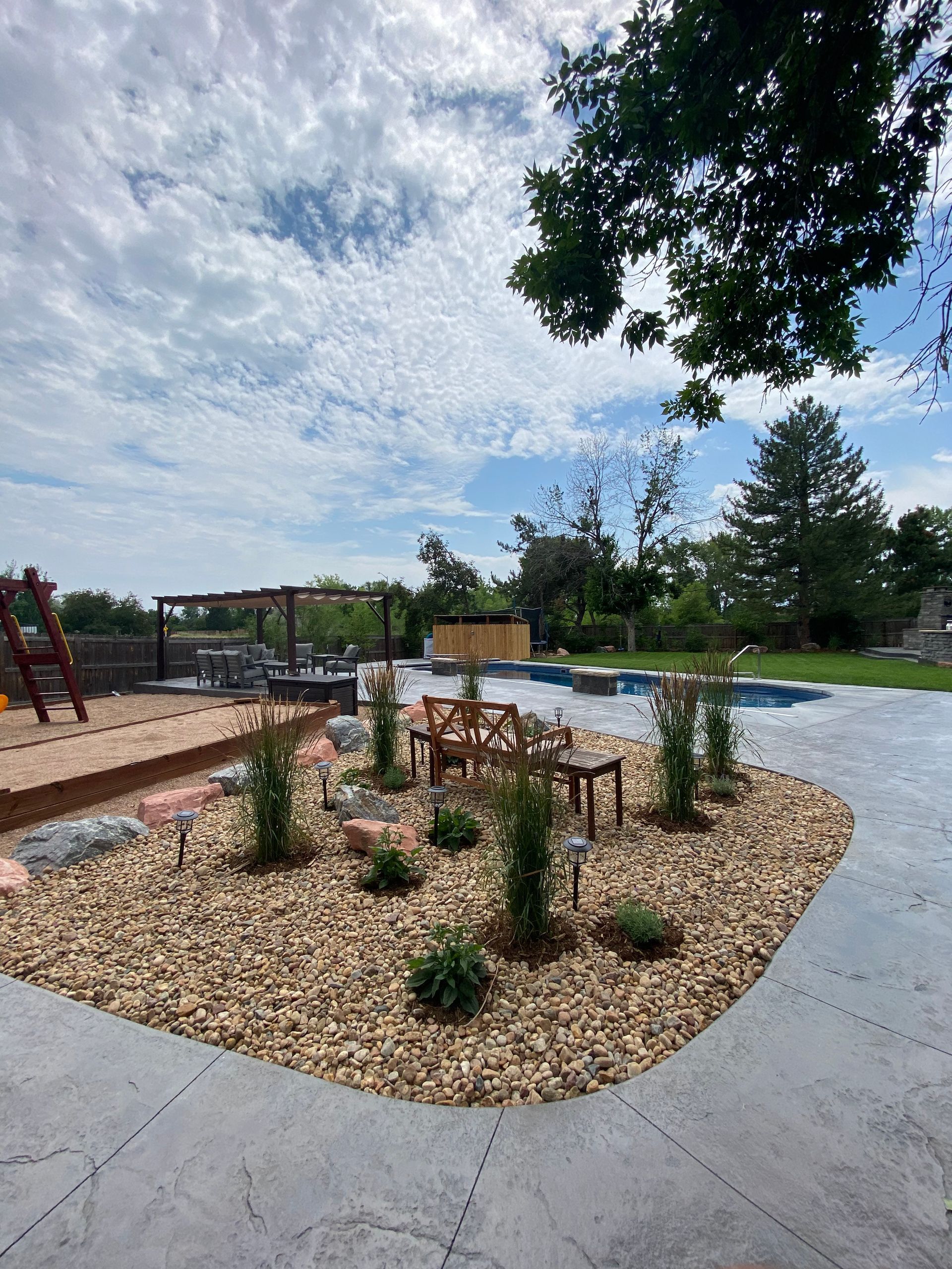 Landscaped yard with rock garden, trees, and patio near a pool under a cloudy sky.