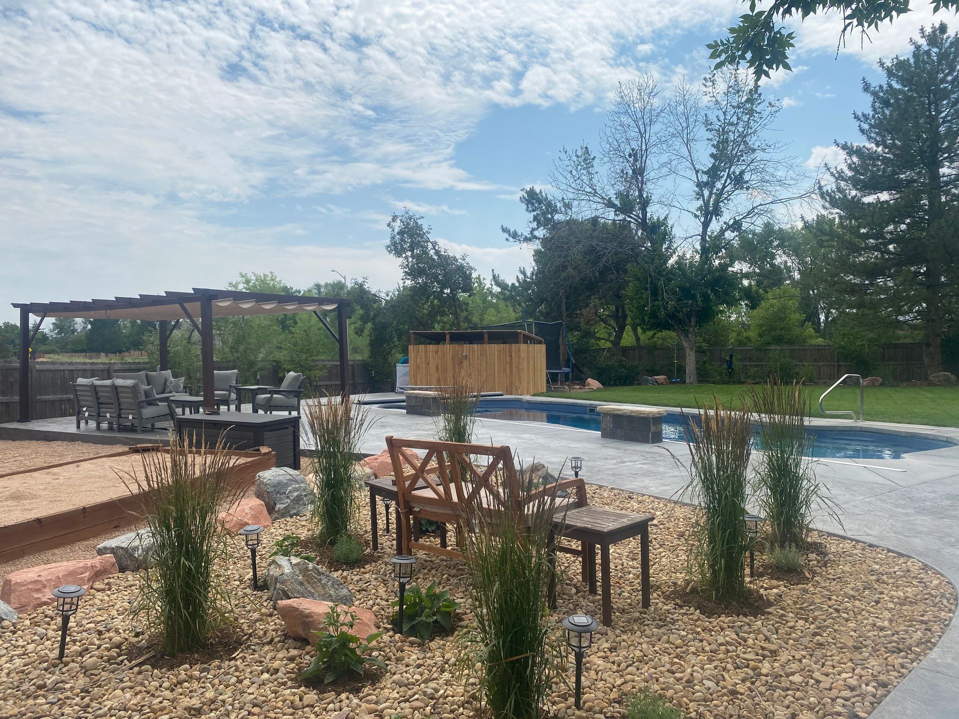 Backyard with pool, hot tub, seating, landscaping, pergola, and blue sky.