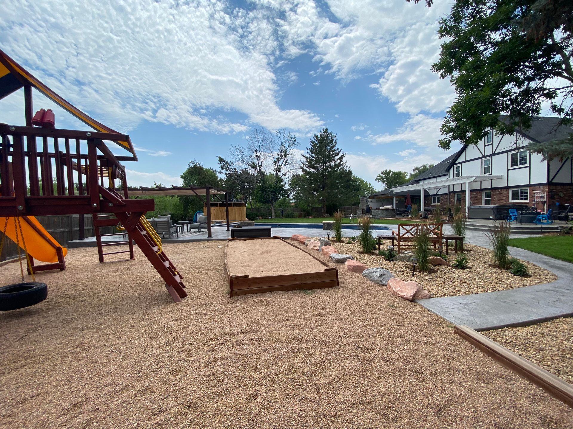Playground with sandbox and wood chips, pool in the background, blue sky with clouds.
