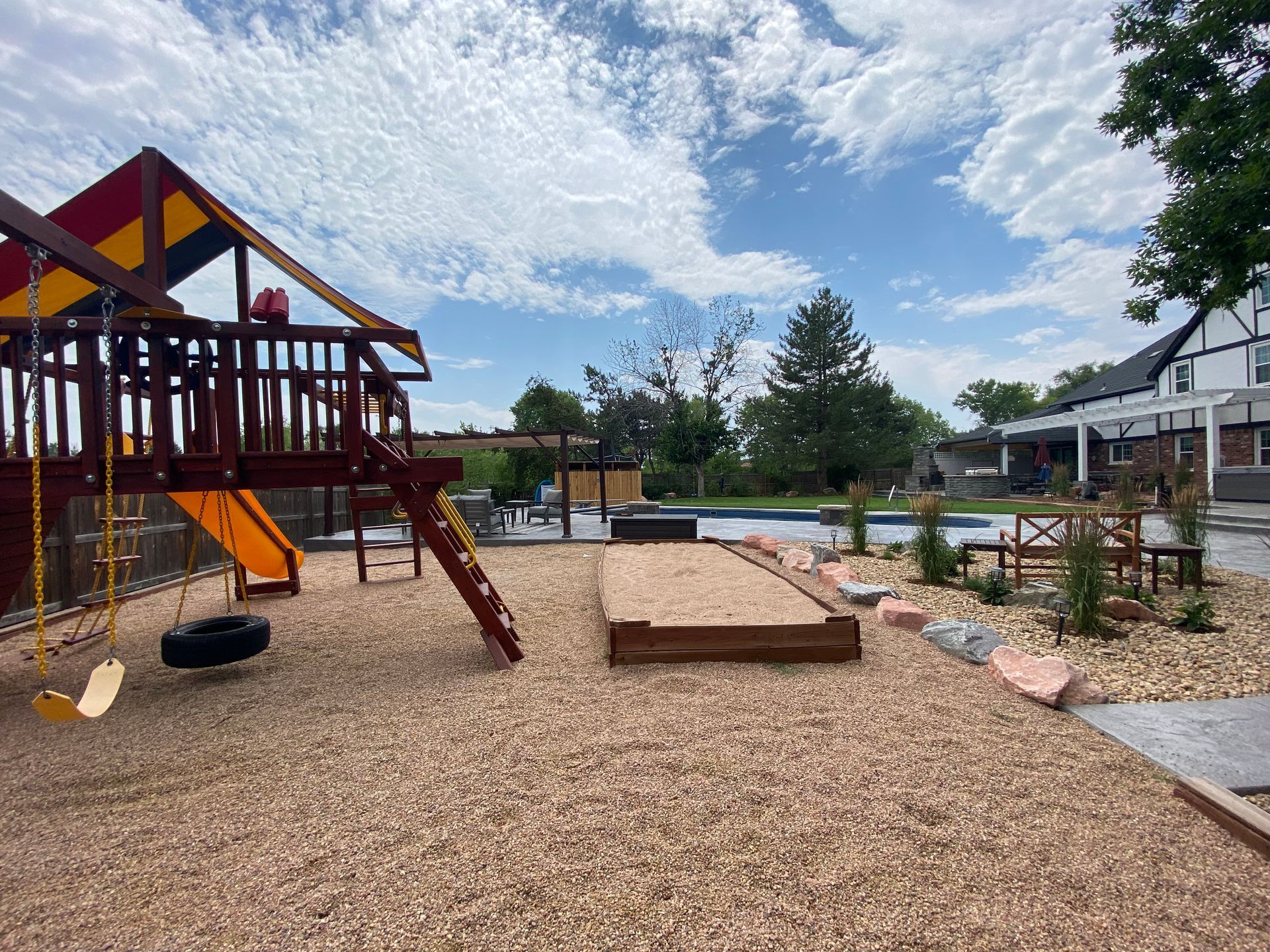 Playground with a slide, swings, and sandbox in a backyard with a pool.