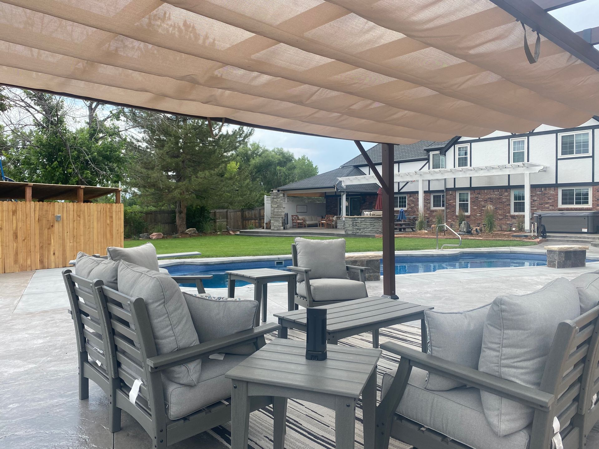 Outdoor patio with gray furniture under a beige shade structure. Pool and house in the background.