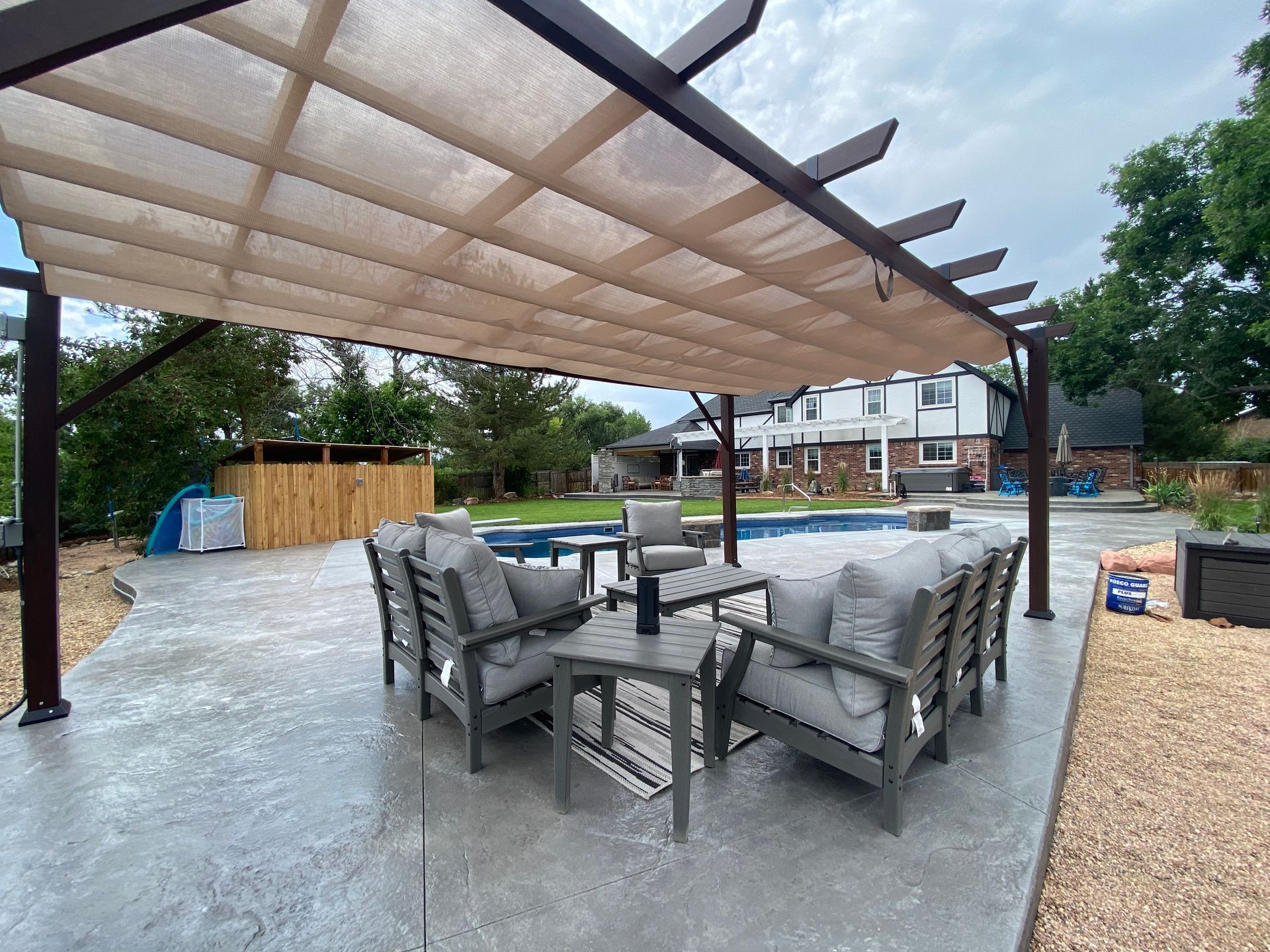 Outdoor seating area under a pergola with beige canopy. Gray furniture on concrete patio by a pool.