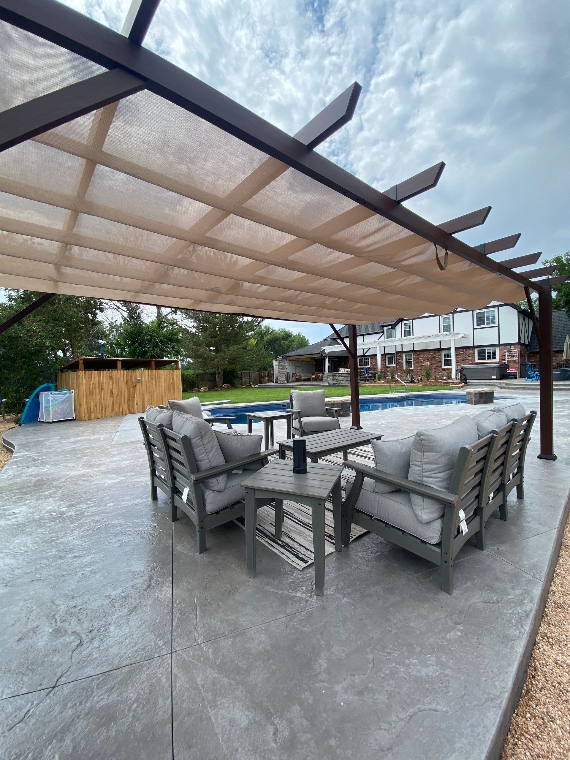 Patio with gray furniture under a brown pergola with a pool in the background. Cloudy sky.