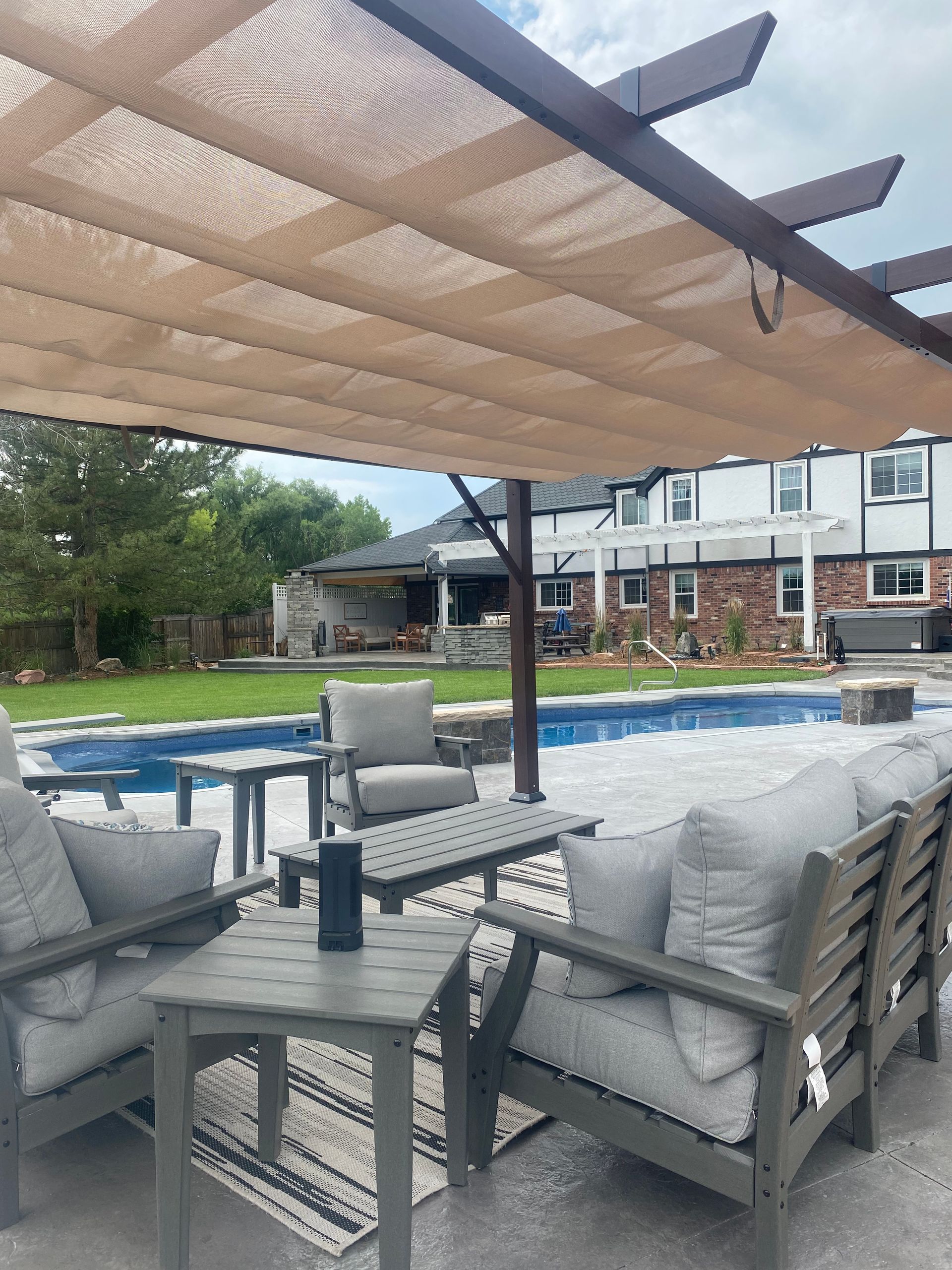 Patio with gray furniture, pool, pergola with shade, and a two-story house in the background.