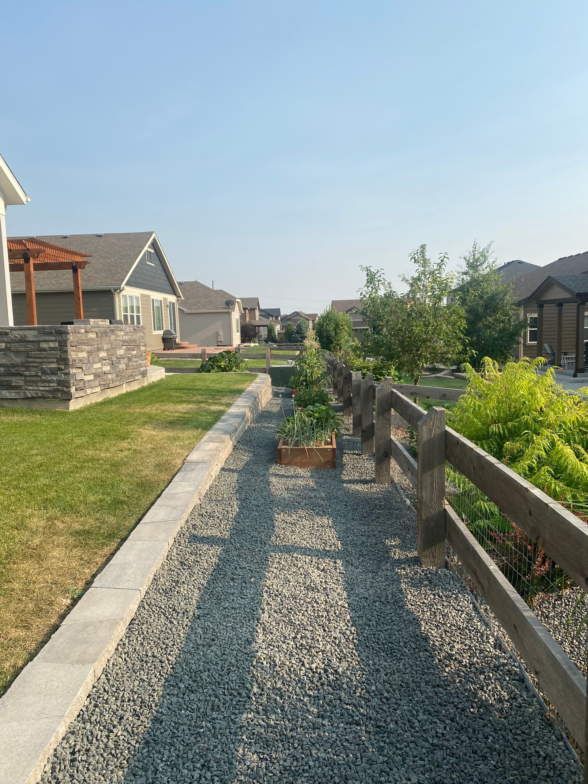 Stone path between grass and wooden fence, leading towards houses under a blue sky.