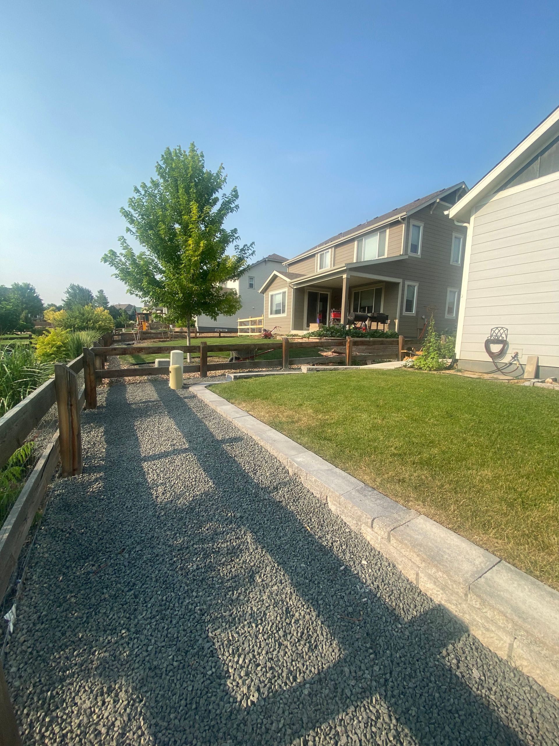 Gravel path next to a wooden fence and grass, leading towards a two-story house with blue sky.