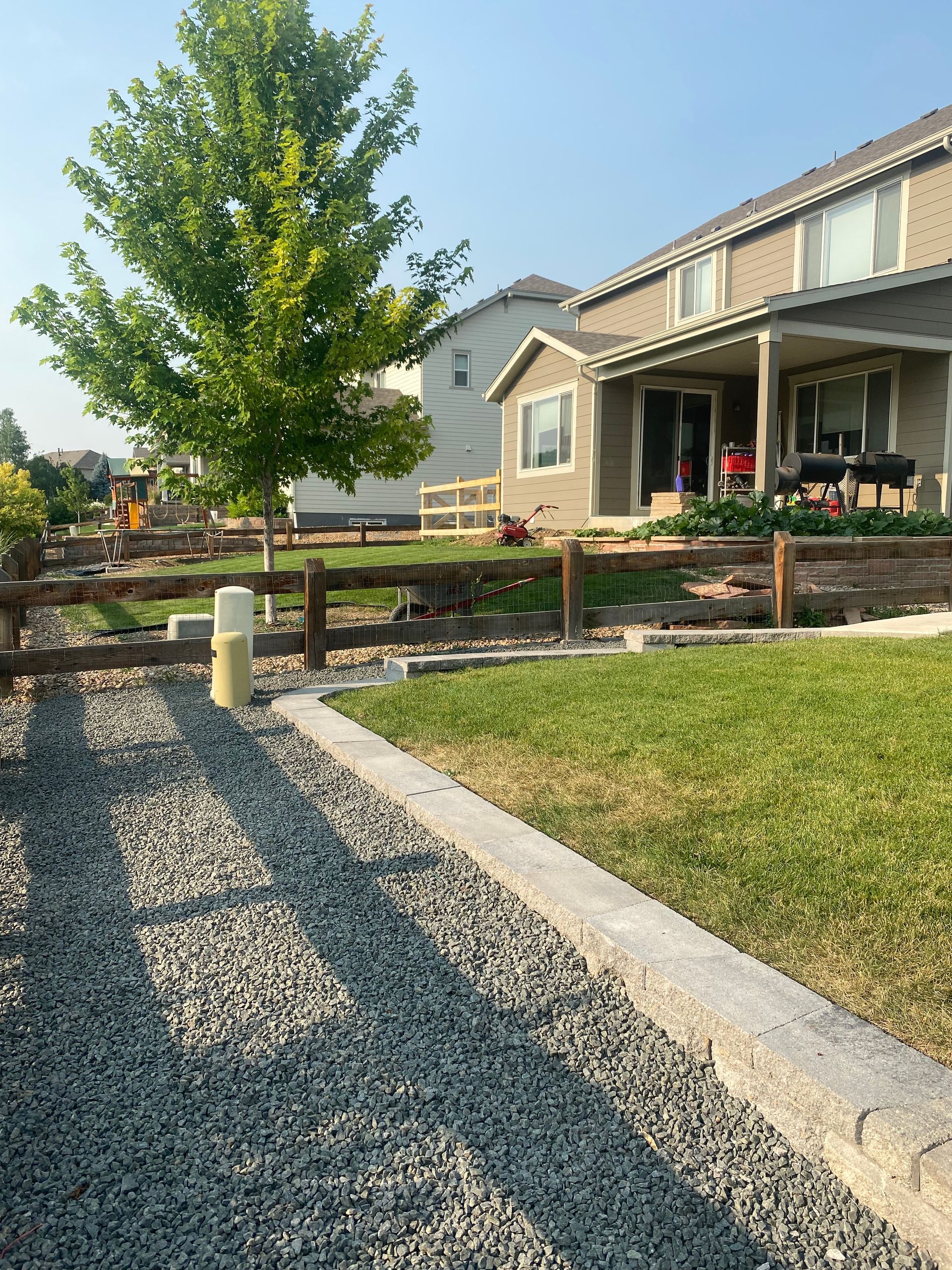 A gravel path leads to a house with a porch and garden, with a wooden fence and a tree on a sunny day.