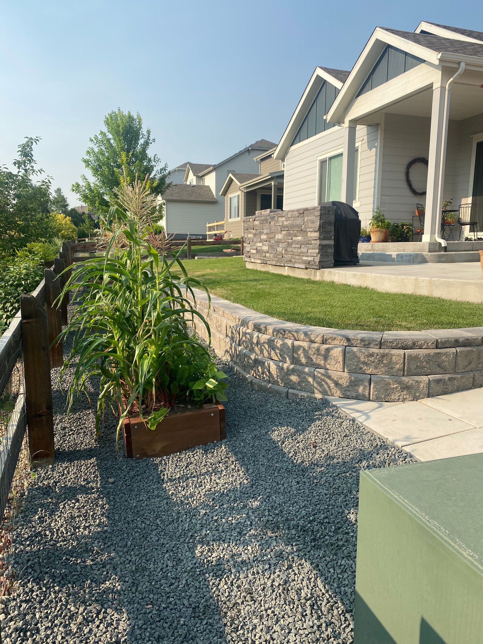 Backyard with raised garden bed, stone retaining wall, and houses in the background. Gravel ground, sunny day.