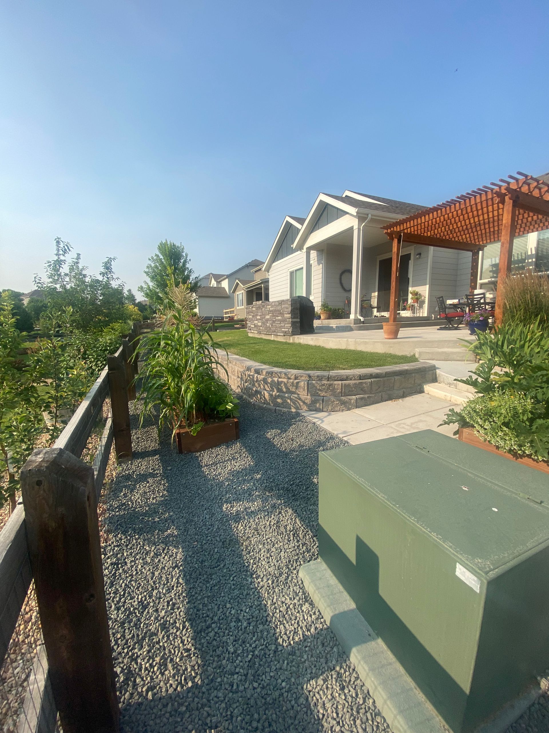 Gravel pathway beside a wooden fence leads to a house with a pergola and raised garden beds, under a blue sky.