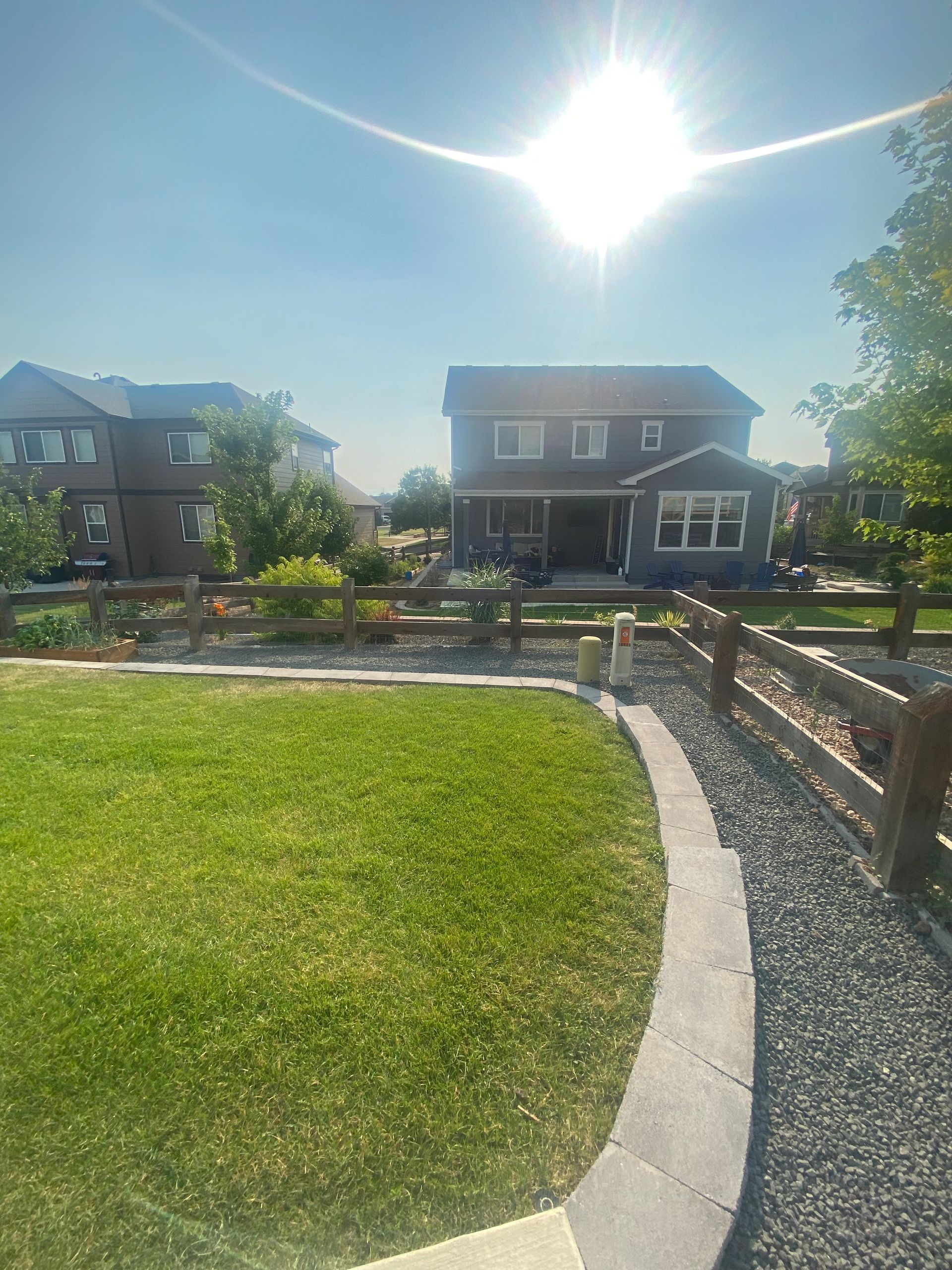 Bright sunny backyard with green grass, stone border, and wooden fence, with houses in the background.