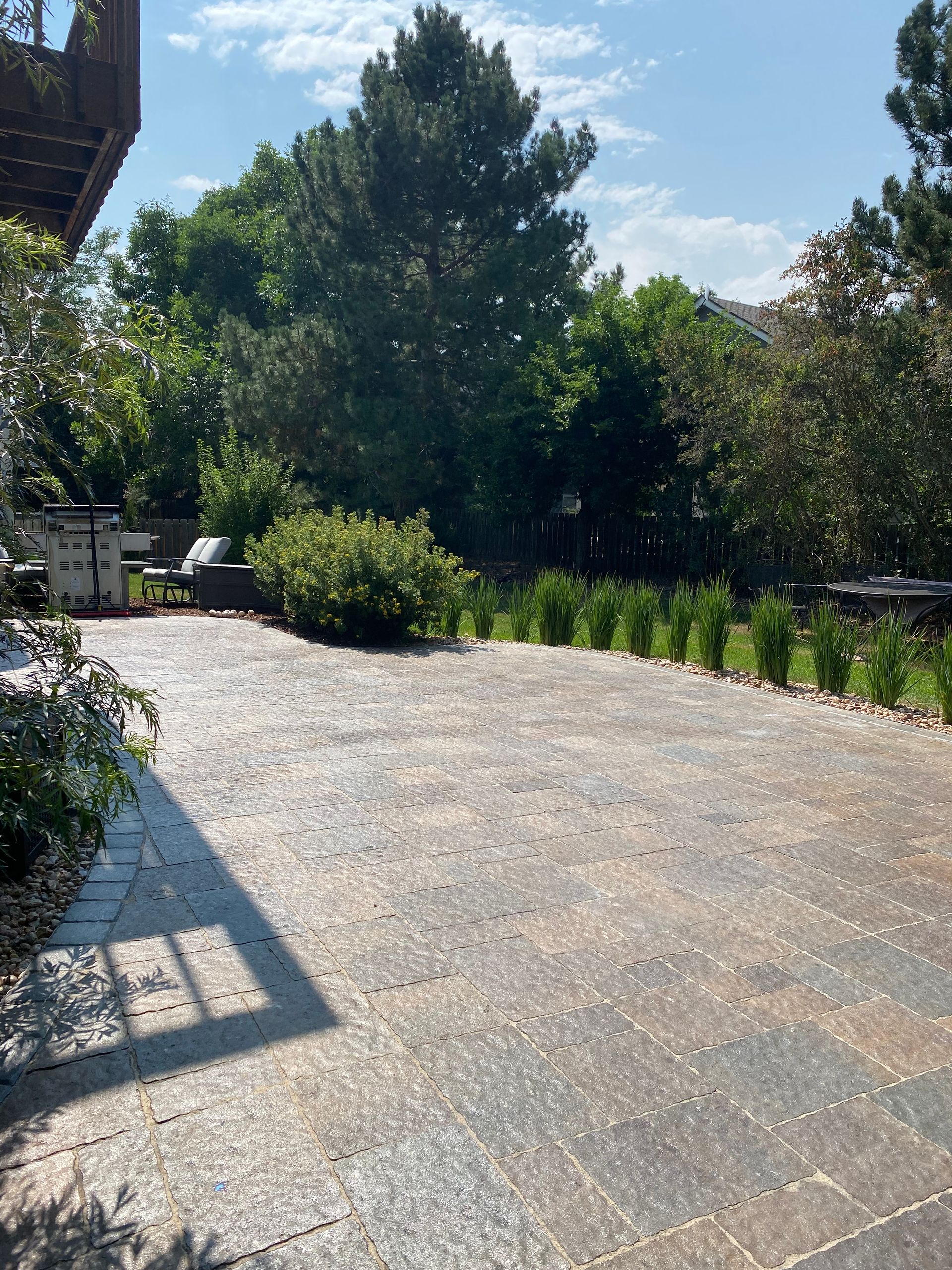 Patio with dark gray pavers, surrounded by greenery and trees under a sunny sky.