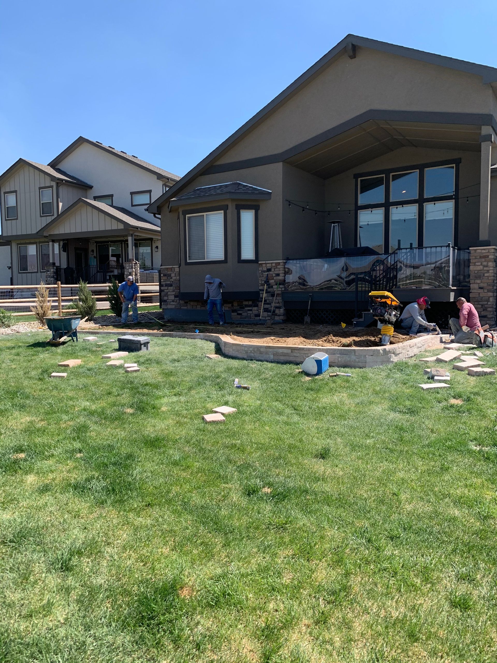 Workers constructing a patio in a backyard with a house, green grass, and blue sky.