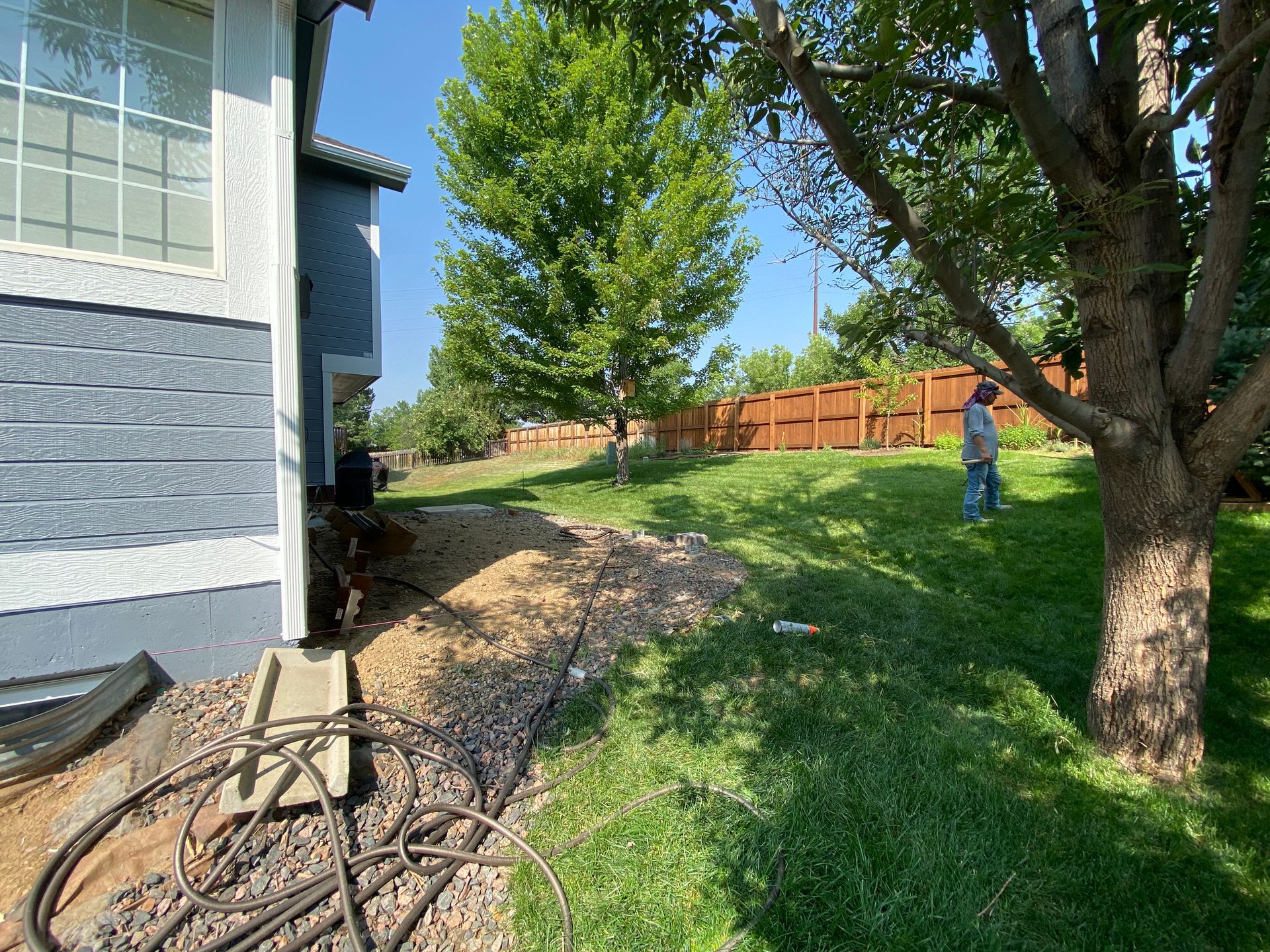 Side view of a blue house with a yard. A person stands near a tree, fence in the distance.
