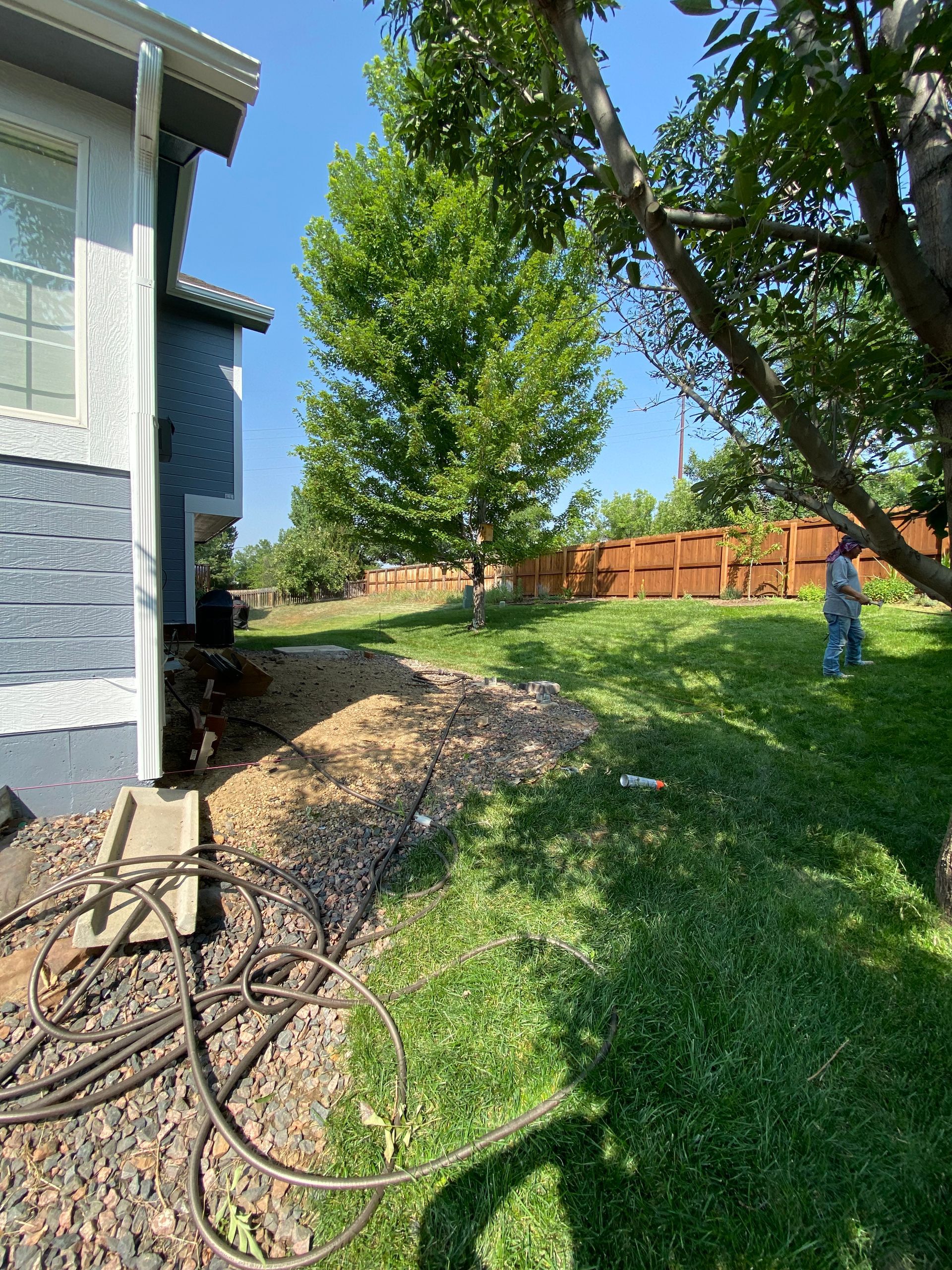 Side yard with a house, gravel path, tree, green grass, and person near wooden fence.