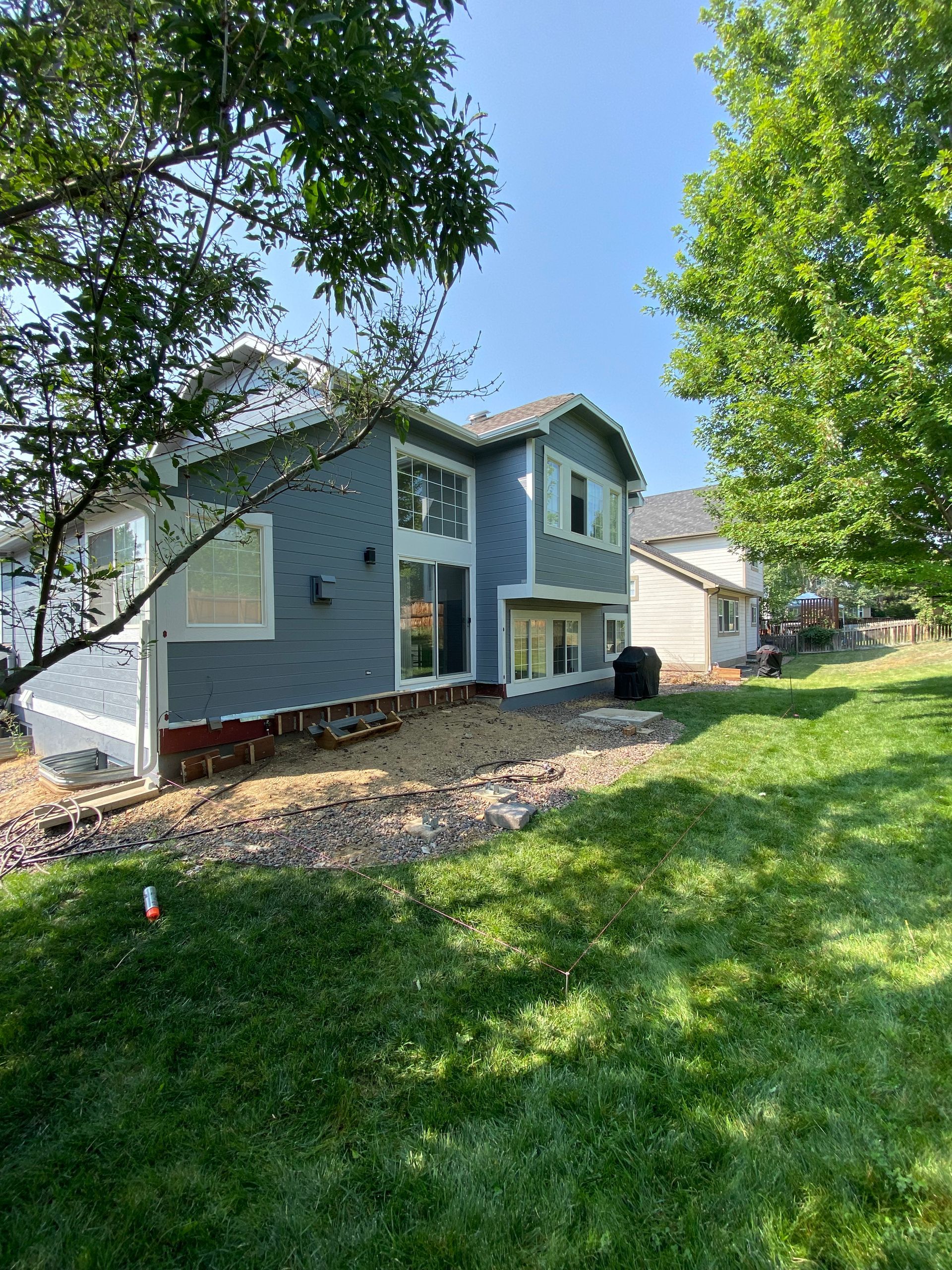 Backyard view of a two-story blue house under construction with a sunny, grassy lawn.