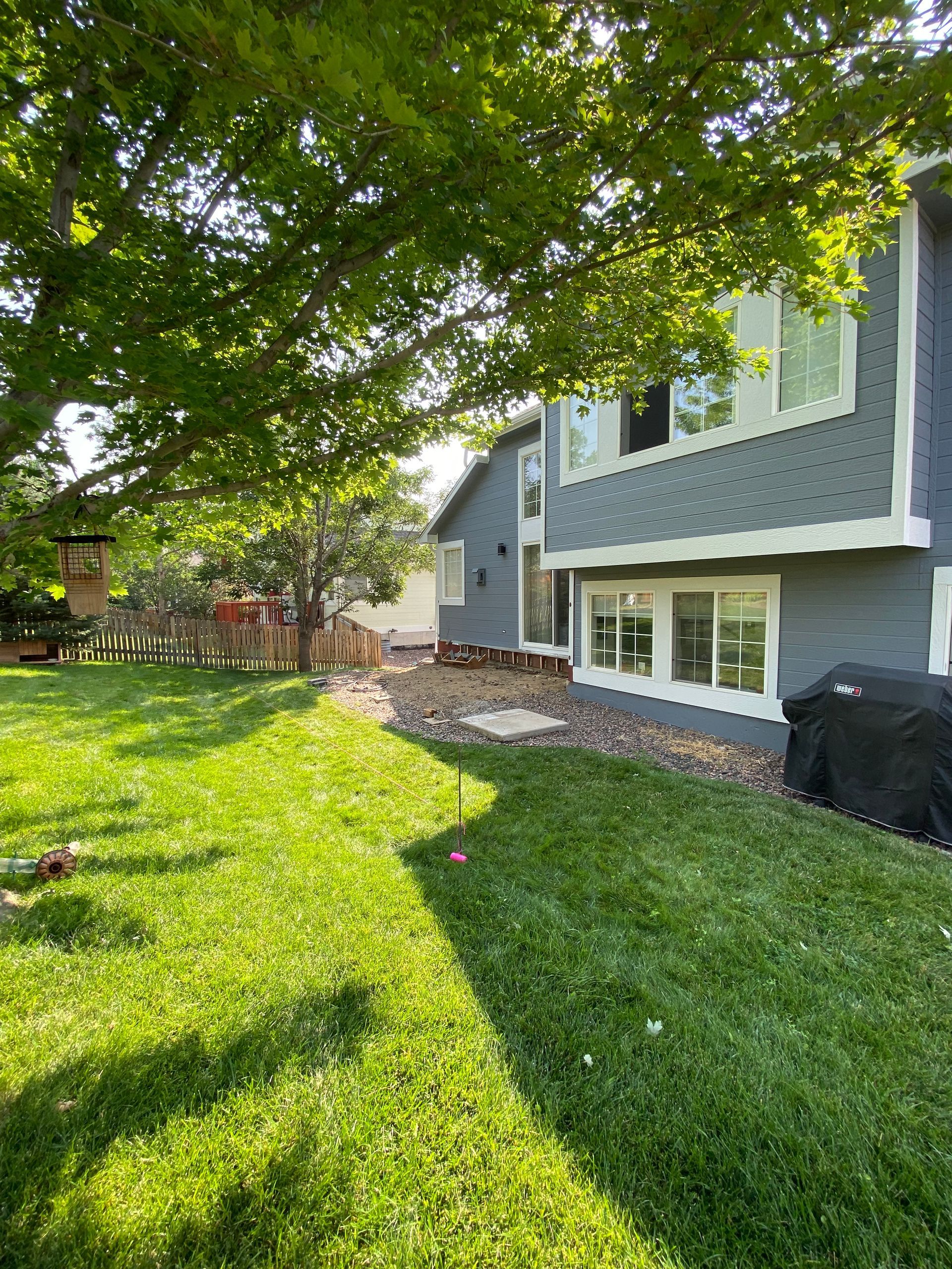 A backyard with green grass, trees, and a gray house with white window frames.
