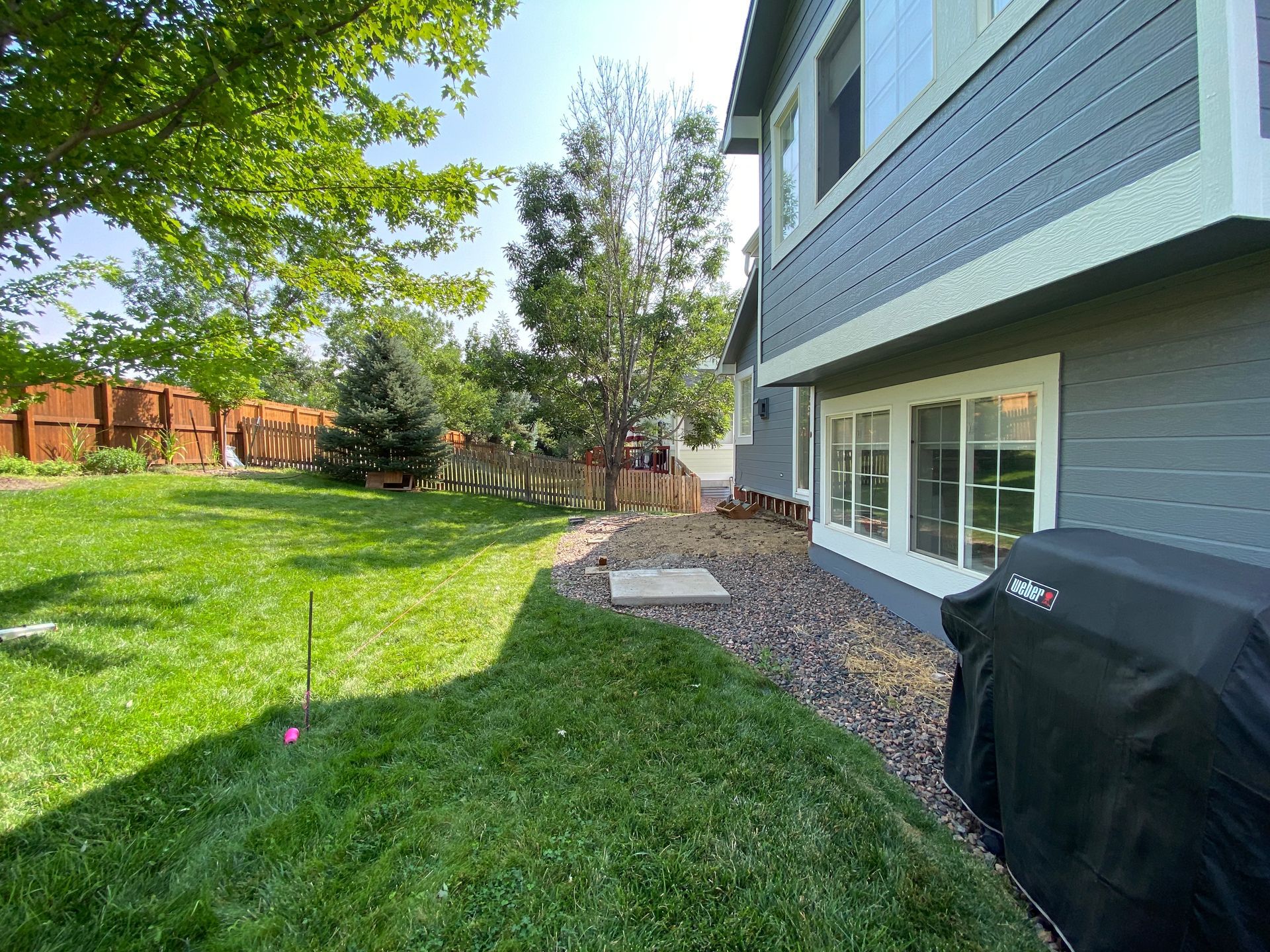 Green grassy backyard with a house, trees, and a grill.