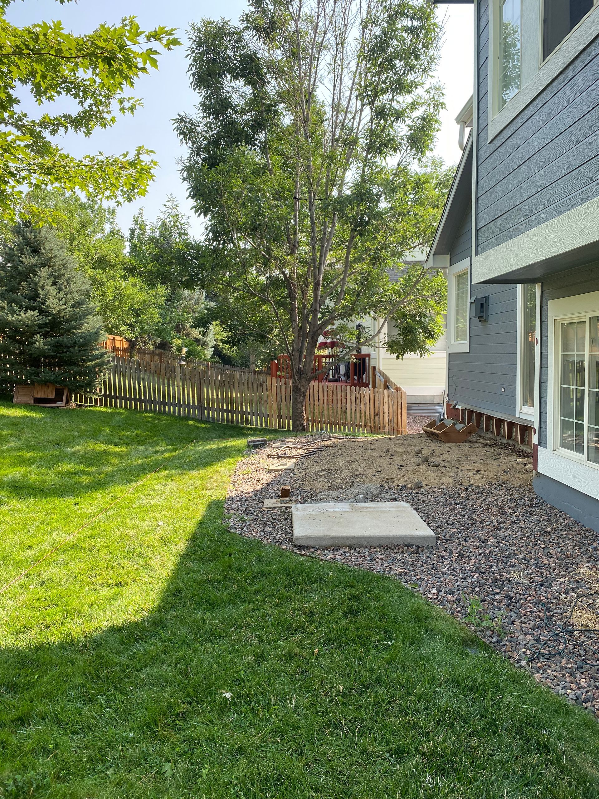 A backyard with green grass, a rock border, and a wooden fence; a house is to the right.