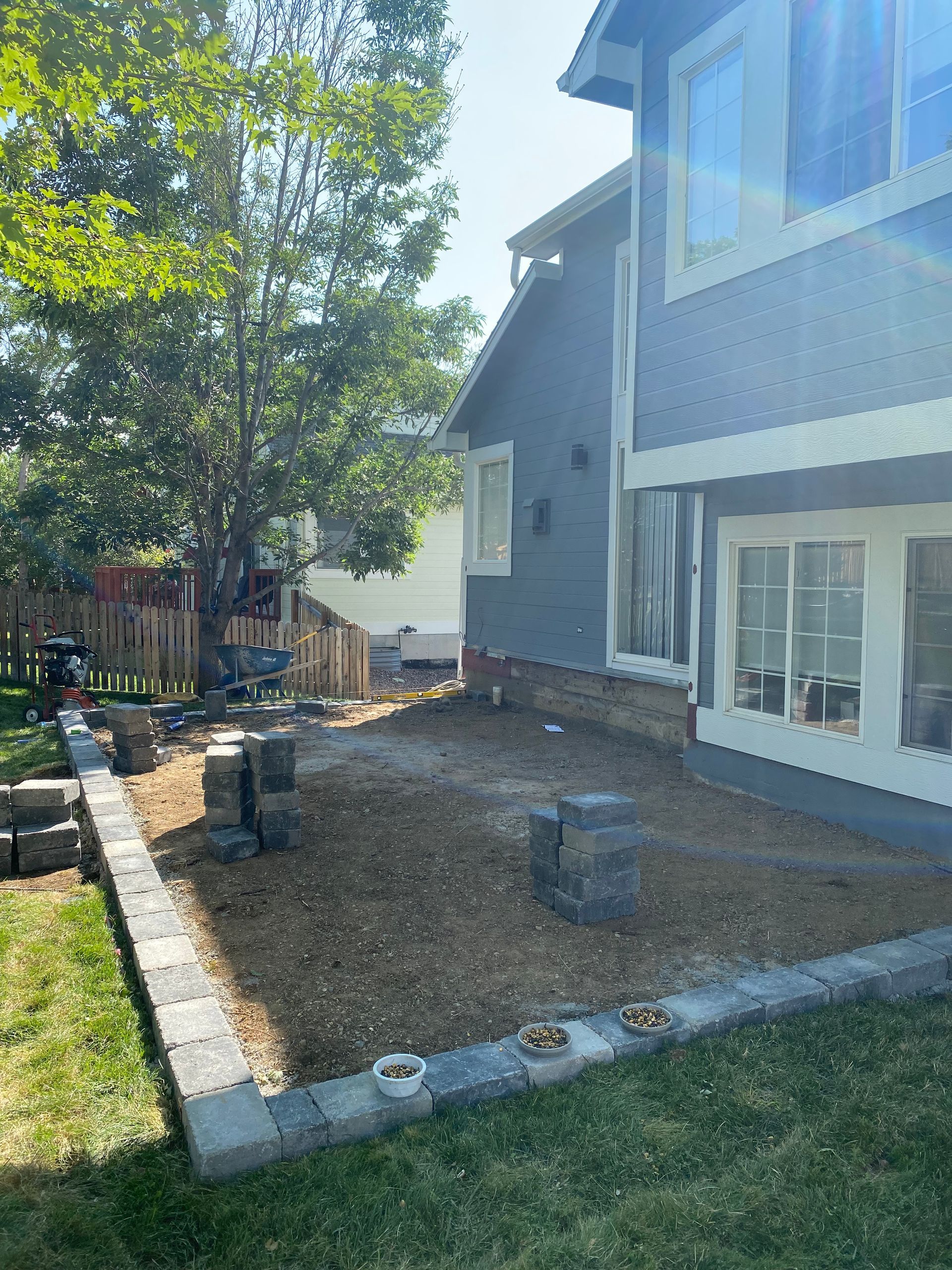 Backyard with construction: gray retaining wall, gravel, house with blue siding, tree, and partial wooden fence.
