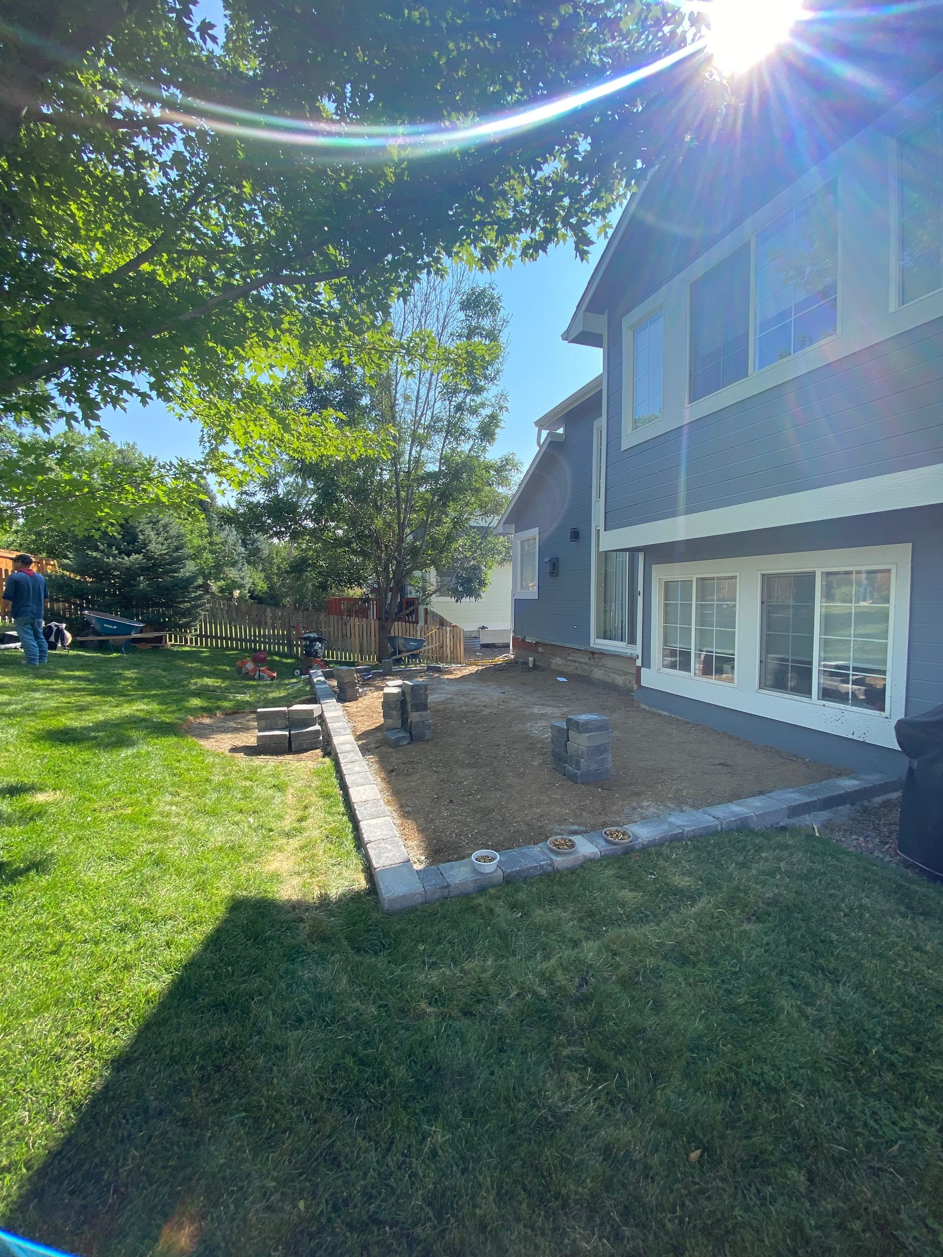 Backyard with gray house, green grass, and a paved patio area with landscaping. Bright sunlight.
