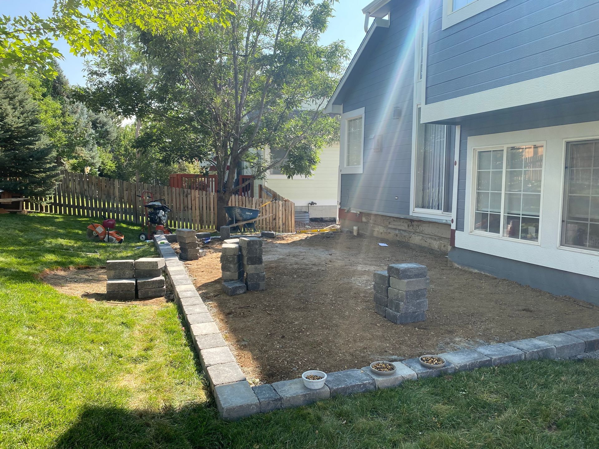Backyard with a stone patio under construction, next to a blue house and a wooden fence.