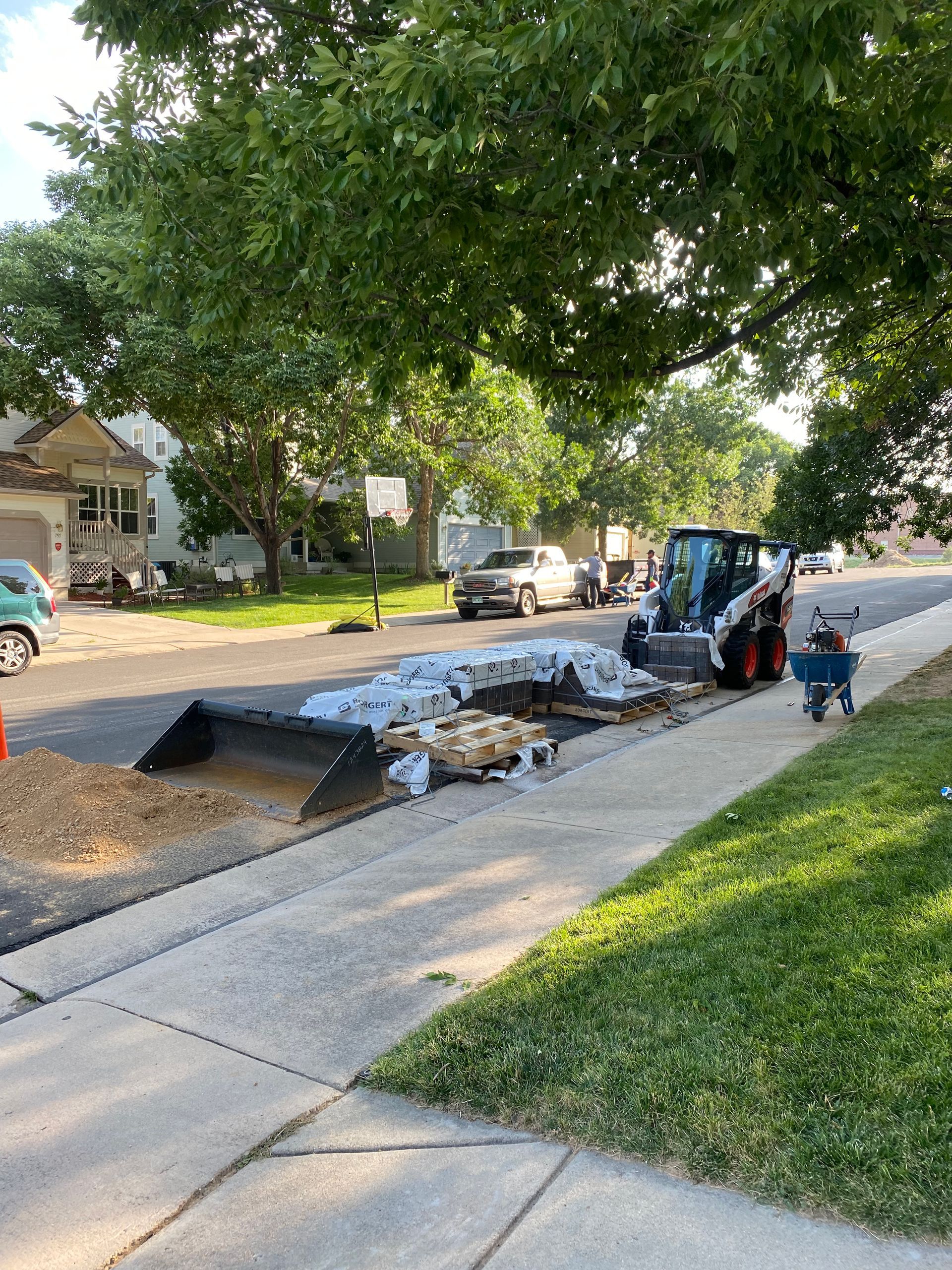 Construction on street. A Bobcat, bags of material, sidewalk, grass, houses, and vehicles are visible.