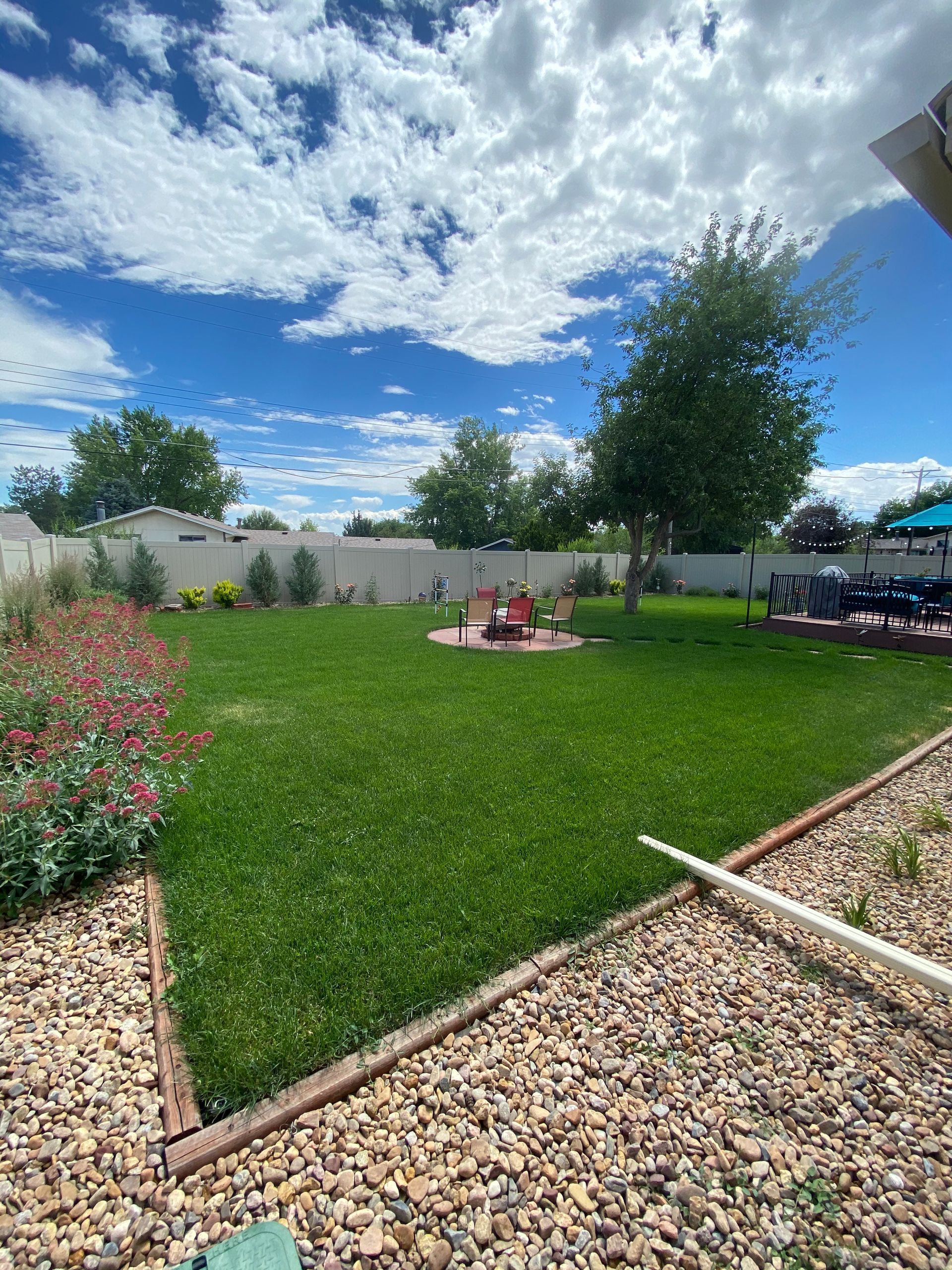Lush green backyard with fire pit, tree, and blue sky with clouds. Brown stone border and red flowers in the front.