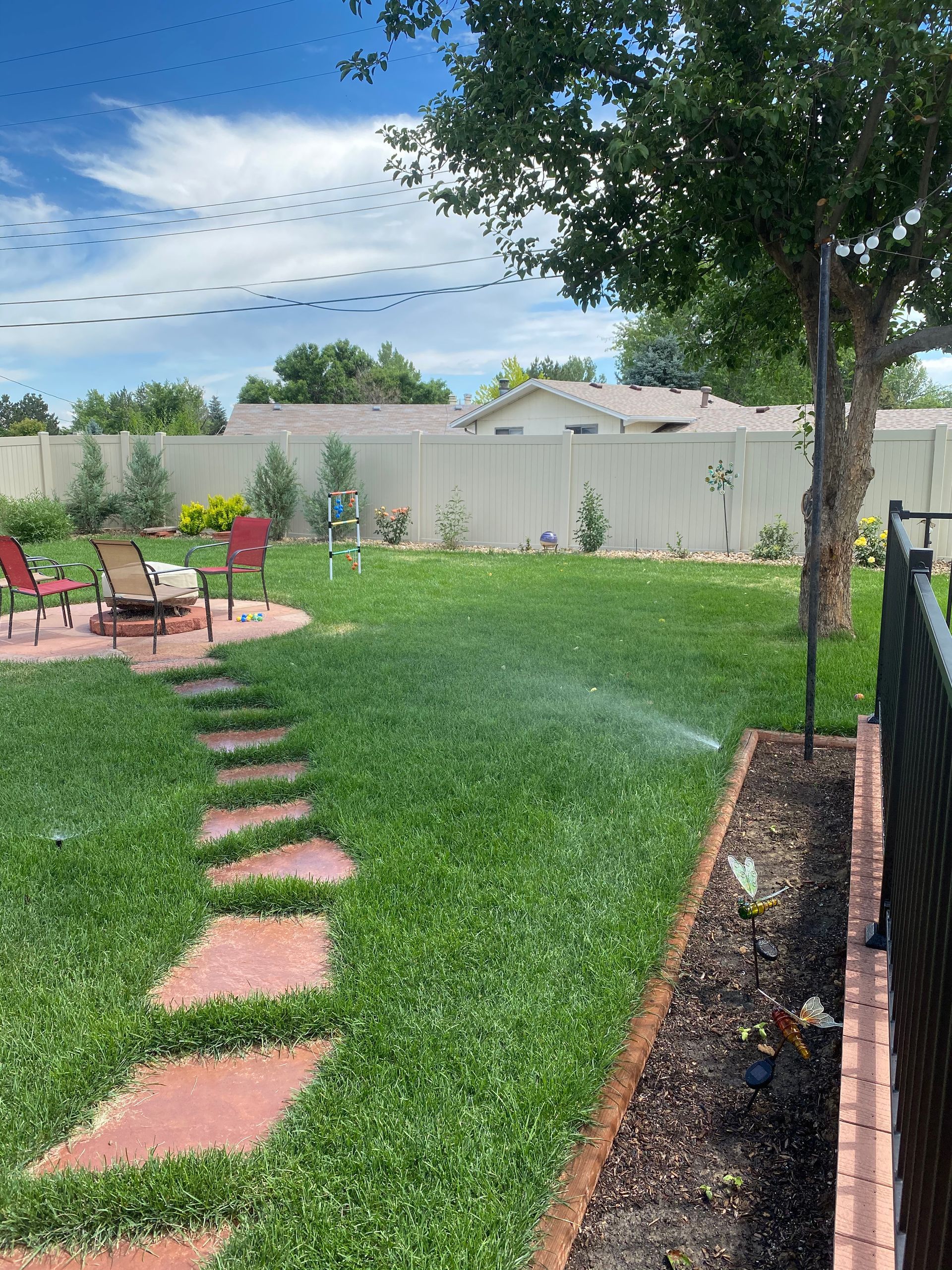 Backyard with green grass, stone path, fire pit, and sprinkler watering lawn. Fence and blue sky in background.