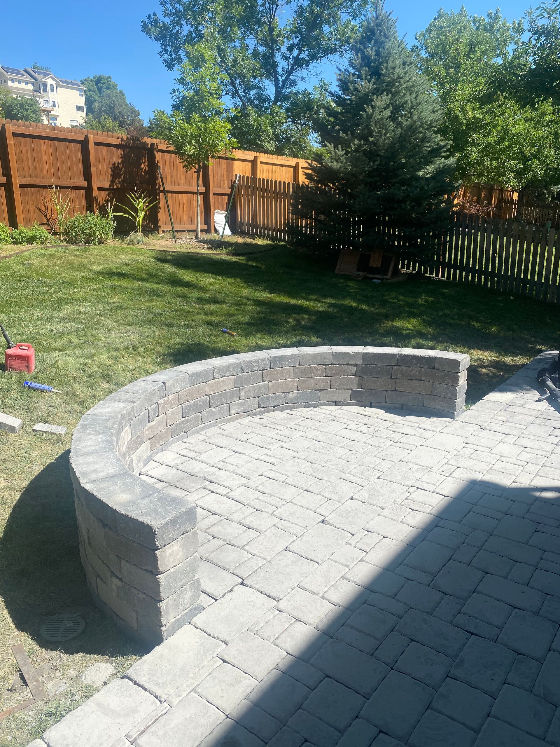 Curved stone patio with matching retaining wall in a backyard with green grass and a wooden fence.