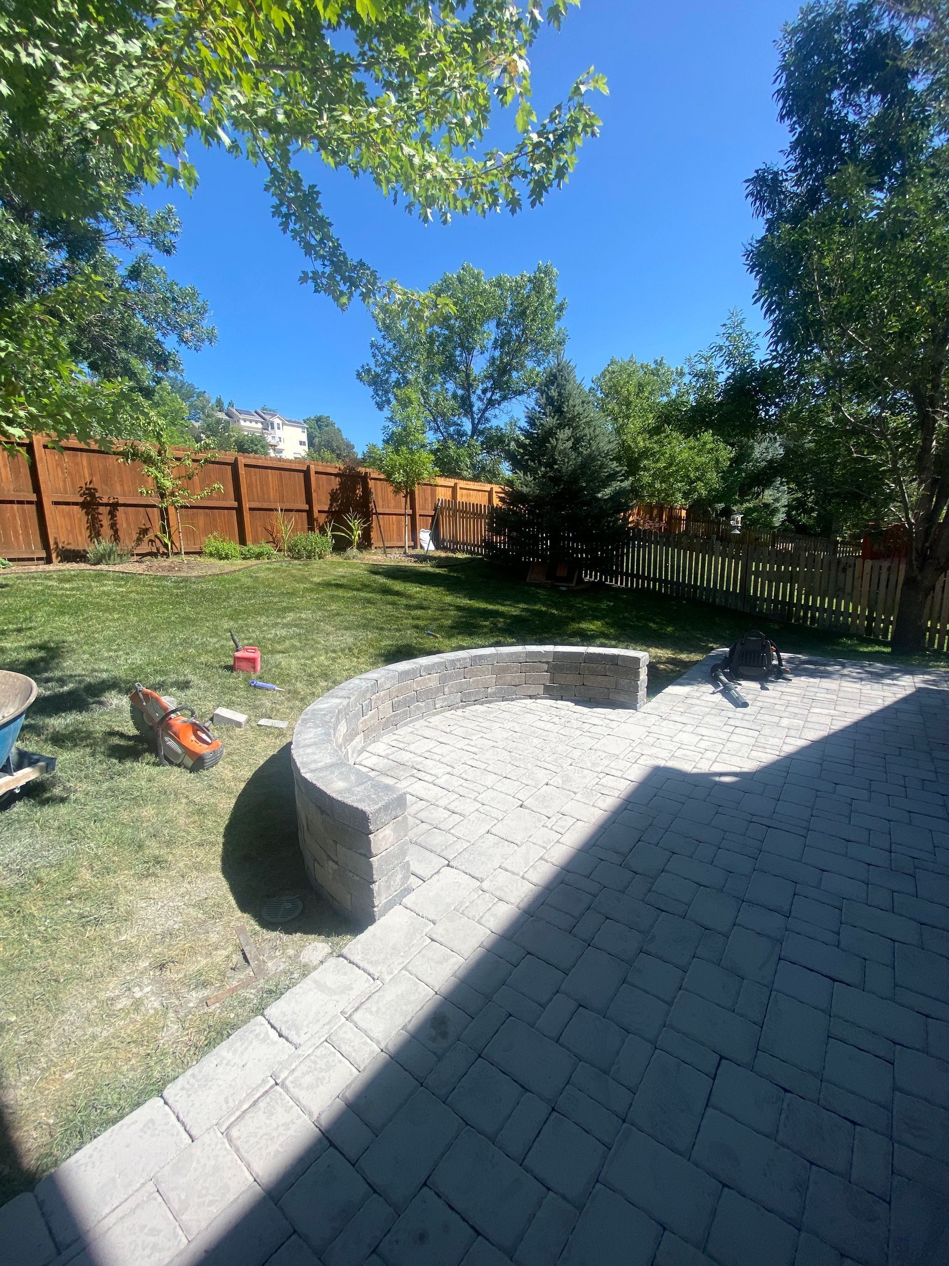 Backyard scene with a stone patio, fire pit, green grass, and wooden fence.