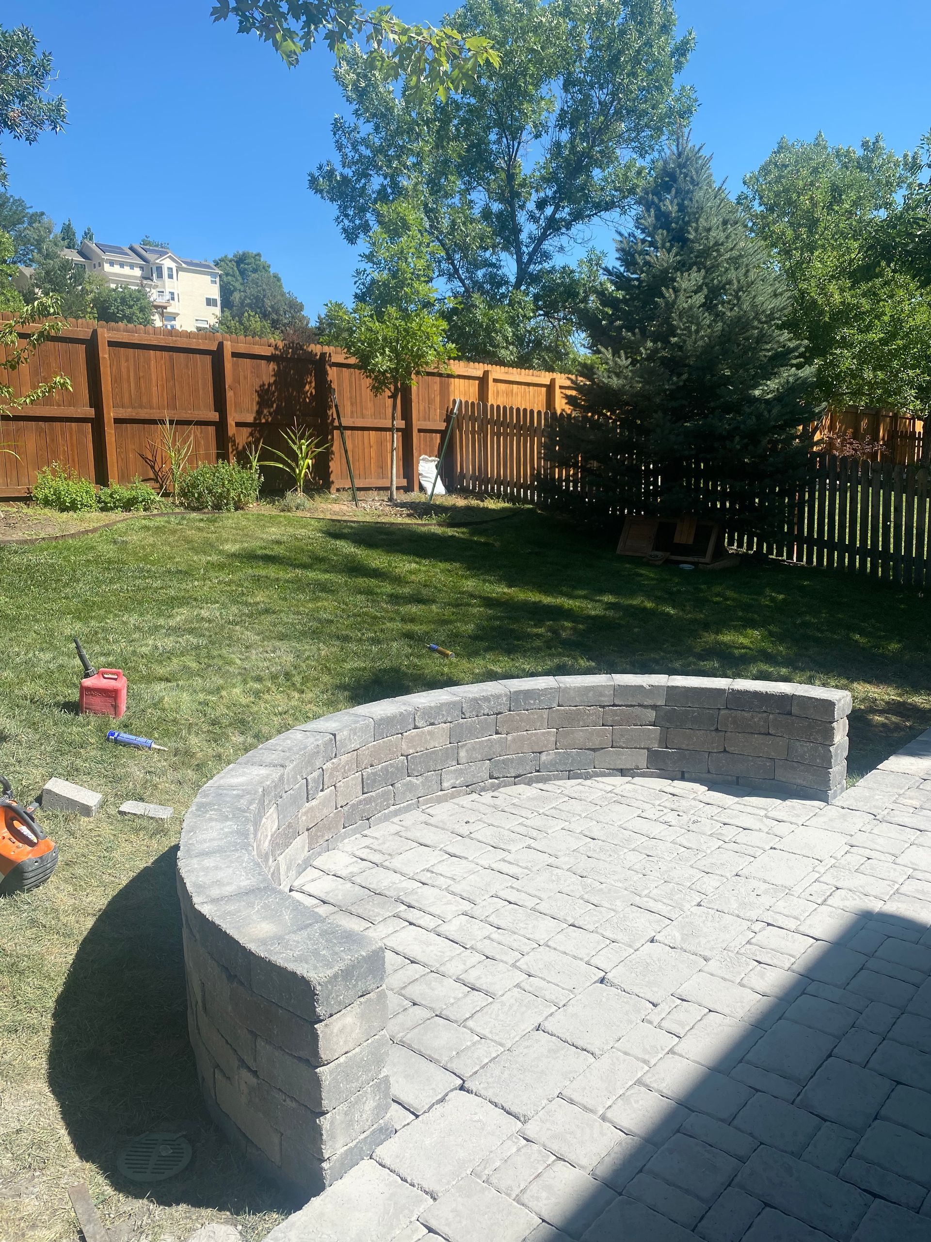 Curved brick patio with retaining wall in a backyard with green grass, a wooden fence, and trees.