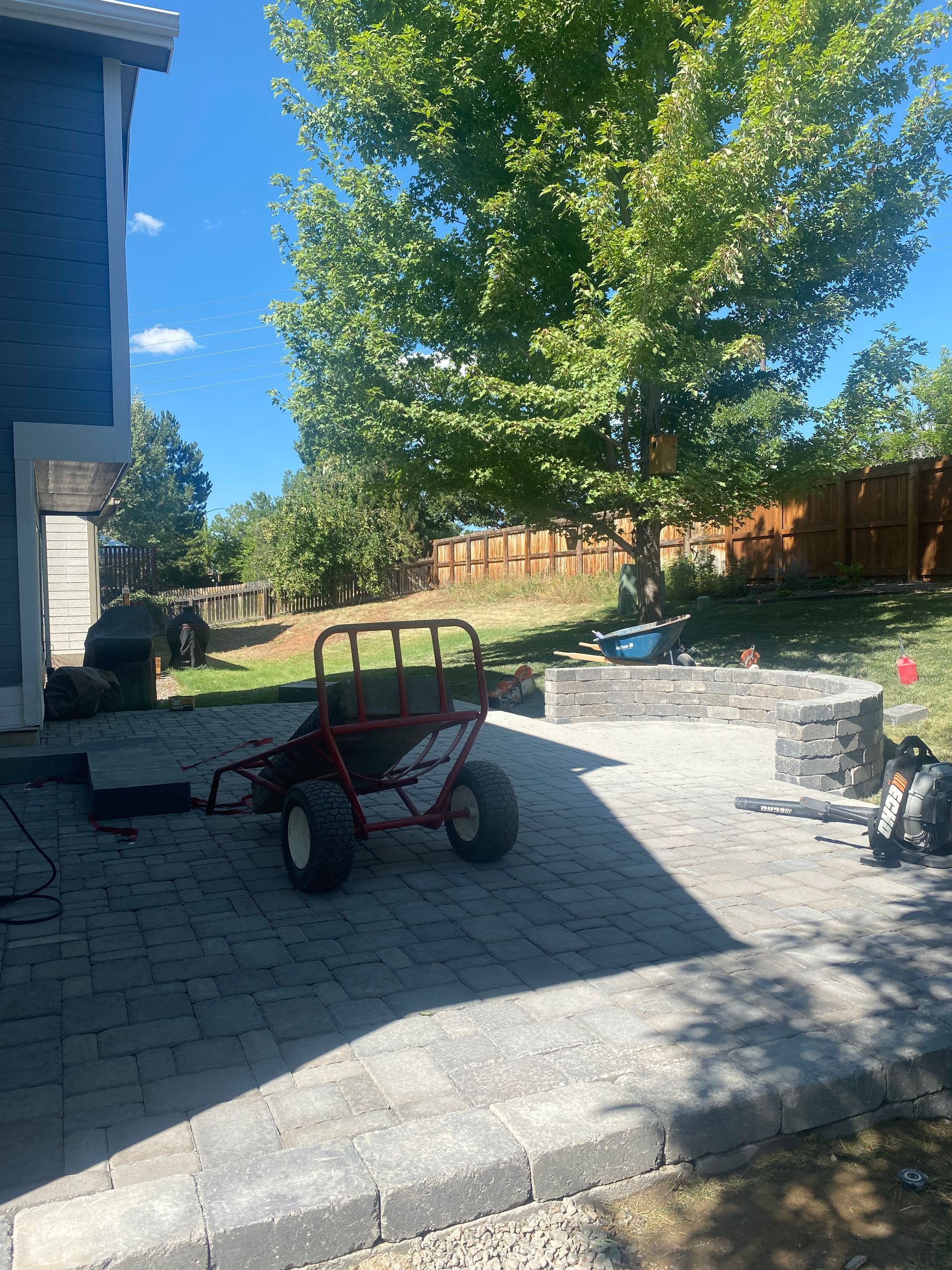 A red utility cart on a paved patio, with a fire pit, large tree, and wooden fence. Sunny day.
