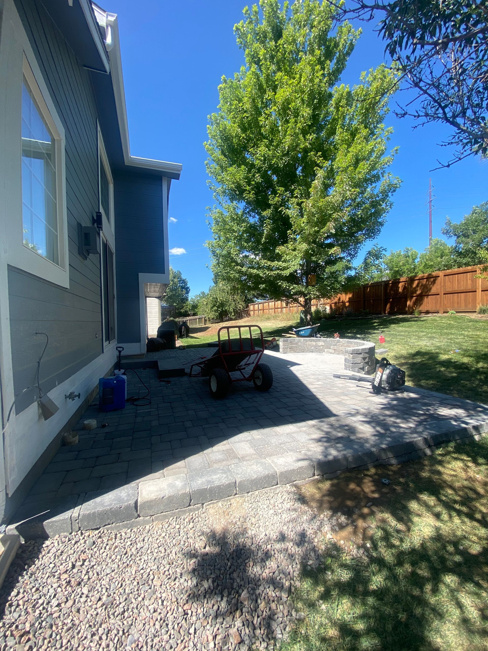 Backyard with gray pavers, gravel path, a tree, and a fire pit near a house.