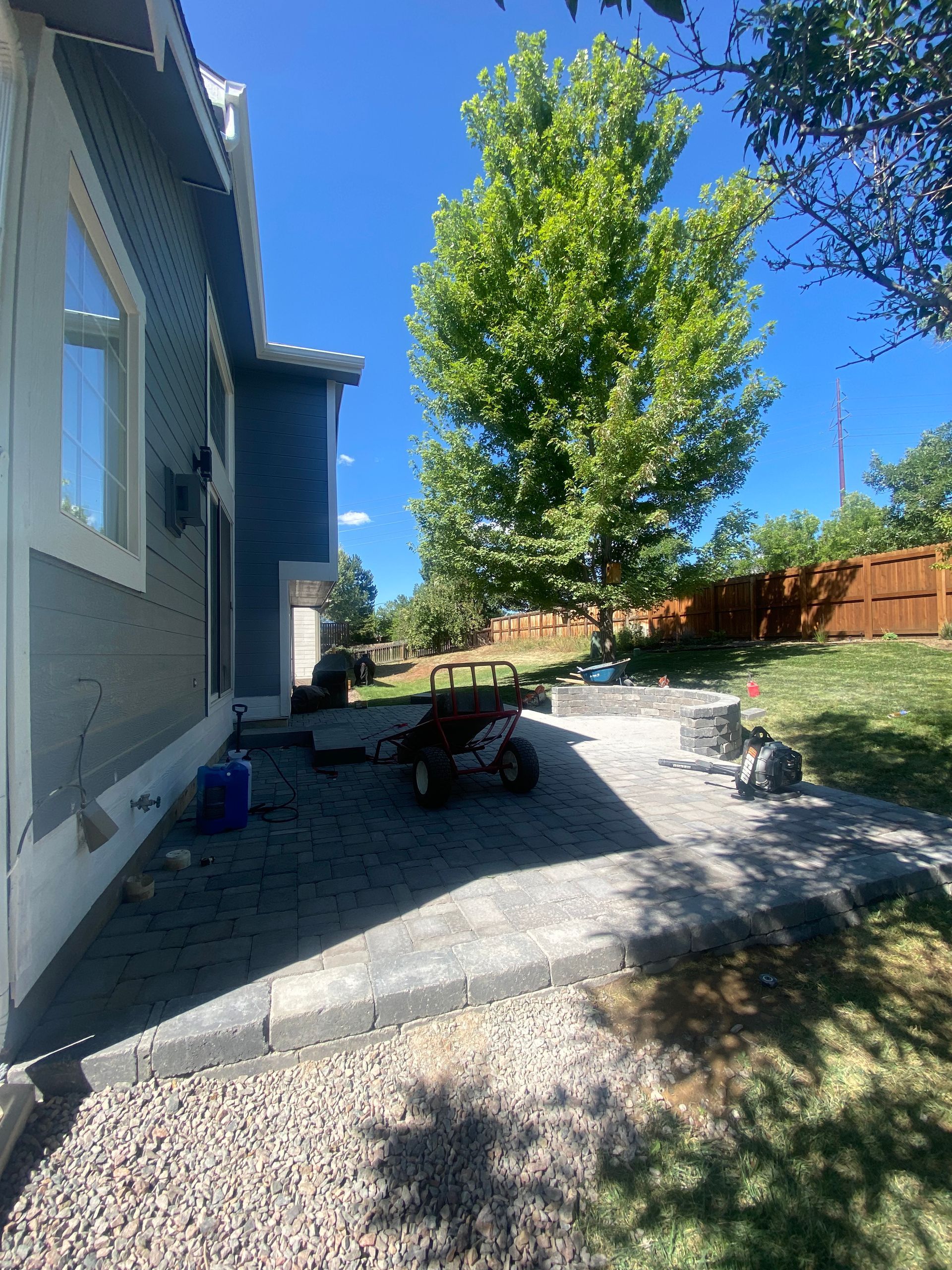 A backyard patio with pavers, a tree, and a small utility cart next to a blue house on a sunny day.