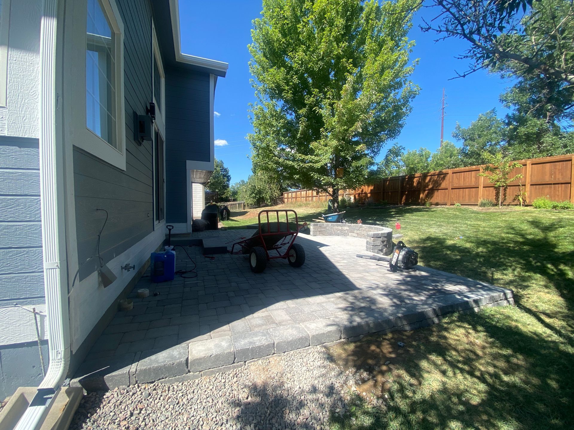 A backyard with a patio, fire pit, and lawn next to a blue house on a sunny day.