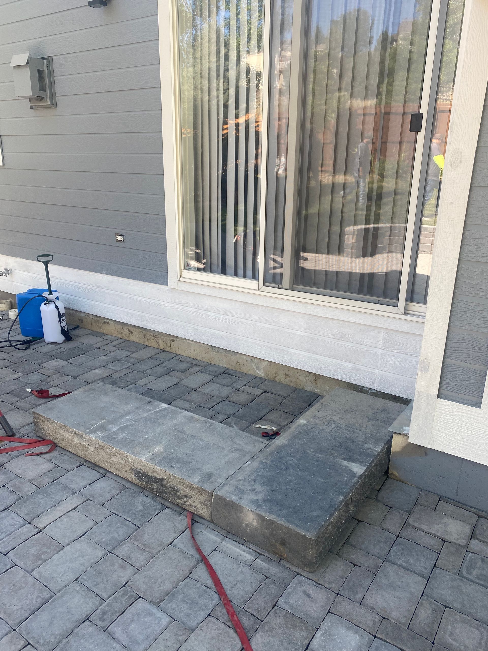 L-shaped gray stone planter on a patio near a sliding glass door. A blue sprayer sits nearby.