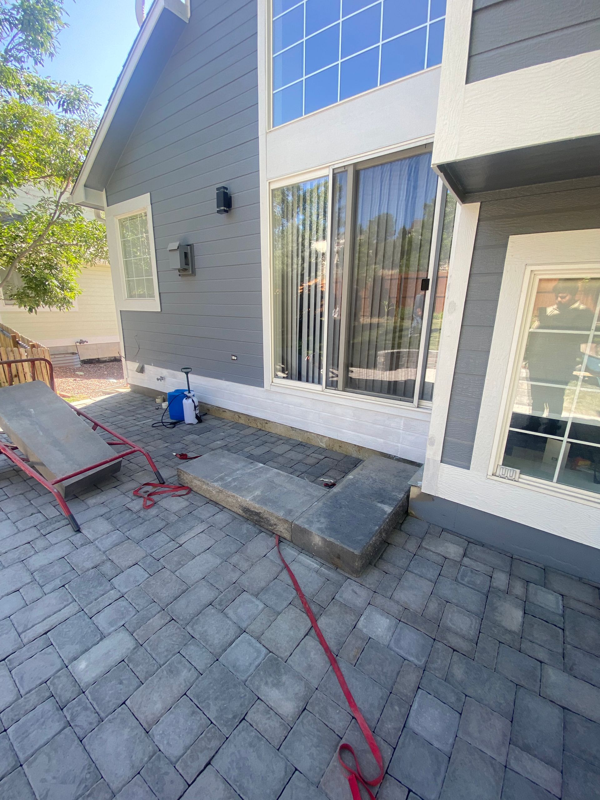 Gray-sided house with a patio under construction, gray pavers, a concrete border, and a red strap.