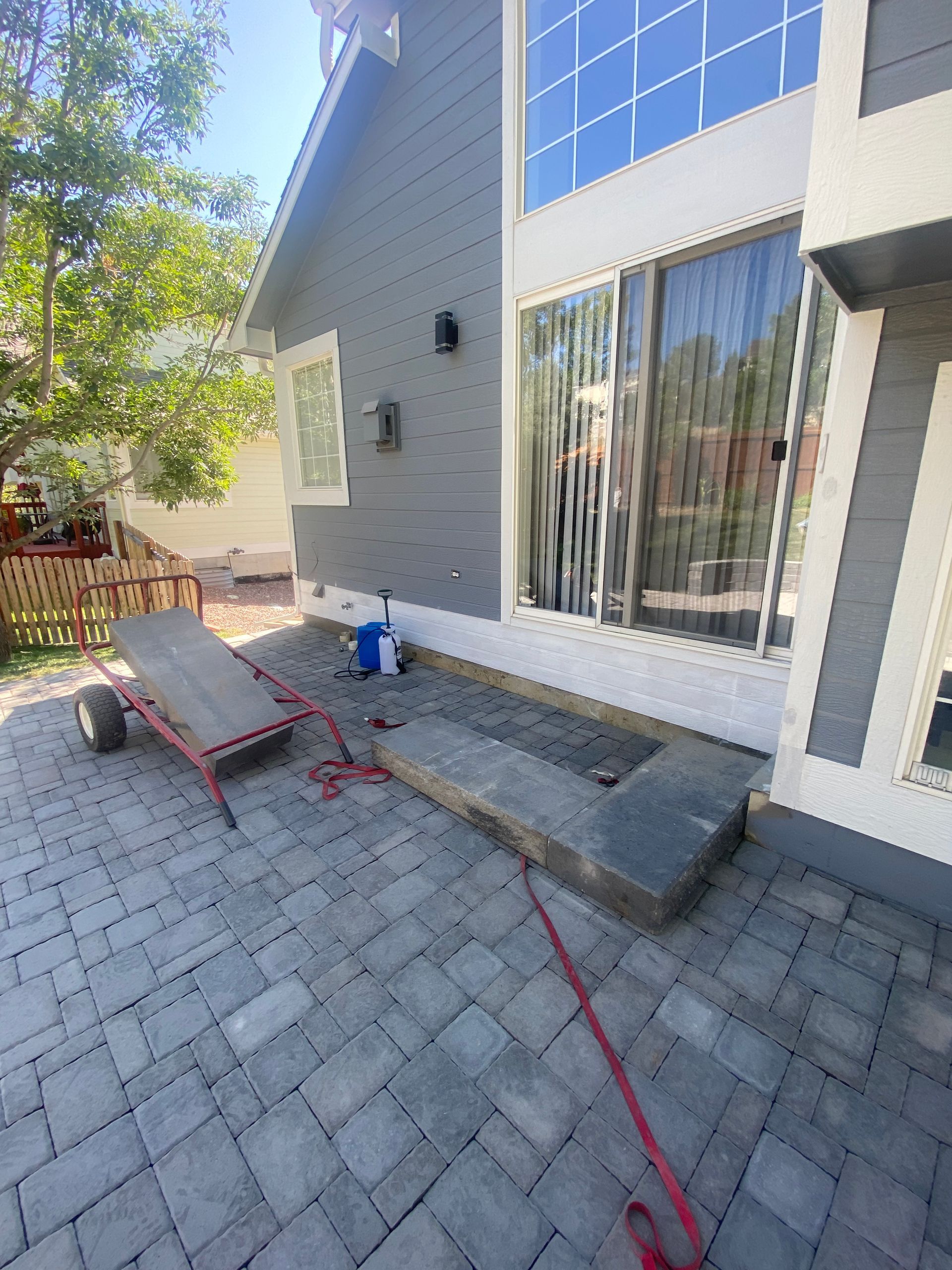Gray house exterior with a paved patio, a rectangular stone feature, and a red garden cart.