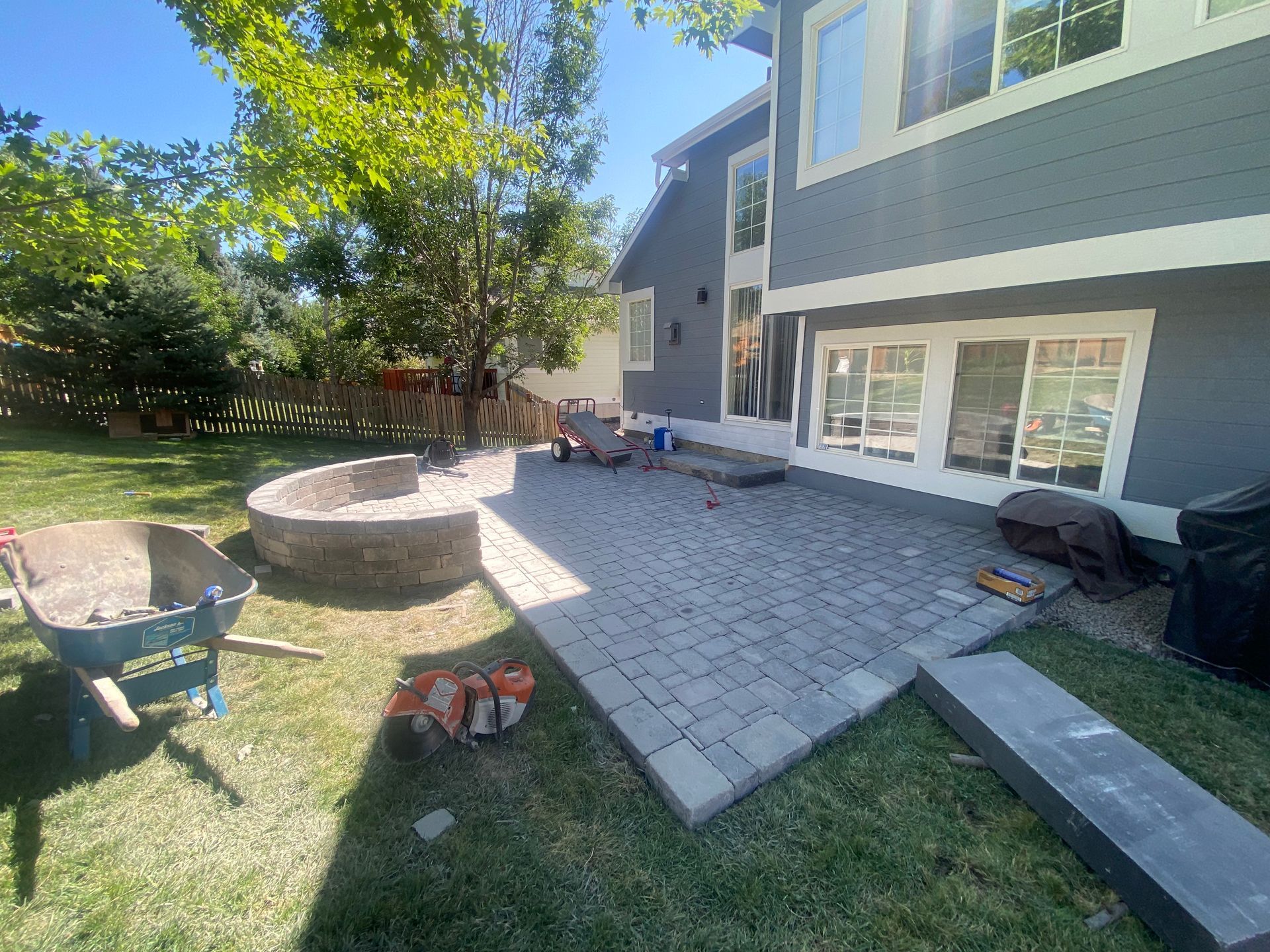 Backyard patio construction with paver bricks, fire pit, and house with blue siding.
